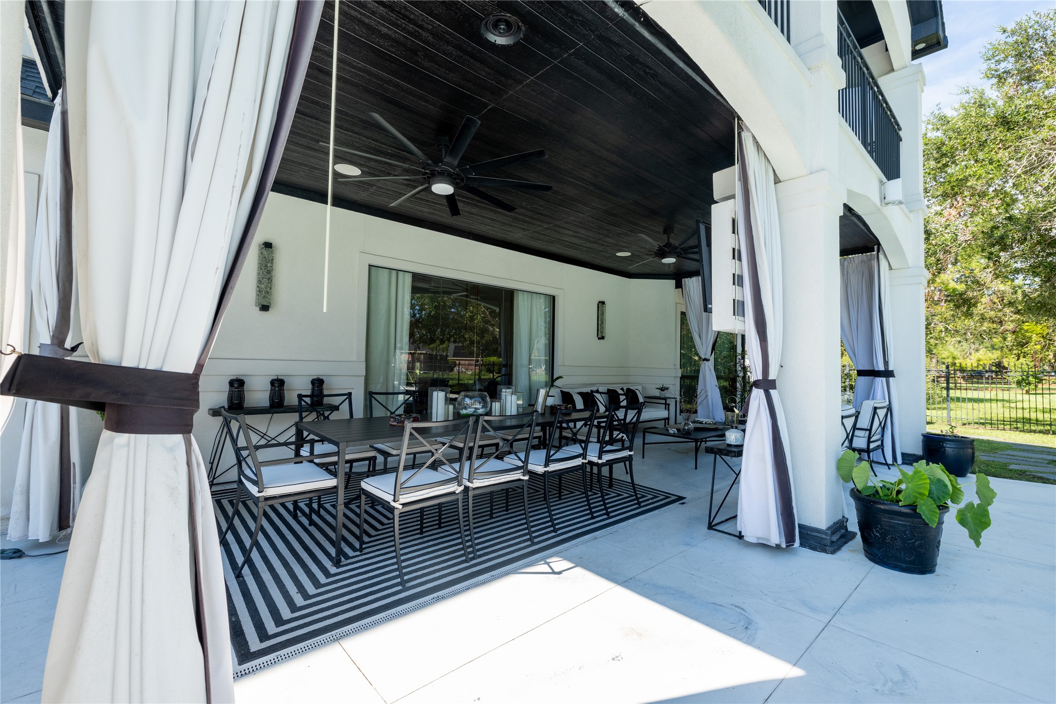 4002 Silver Ridge Boulevard Missouri City, TX 77459 - Photo 41 of 50 This is a spacious covered patio area featuring elegant outdoor dining furniture, cooling ceiling fans, and flowing curtains that add a touch of privacy and sophistication. The design incorporates a contemporary black and white color scheme, offering a stylish space for relaxation and entertainment.