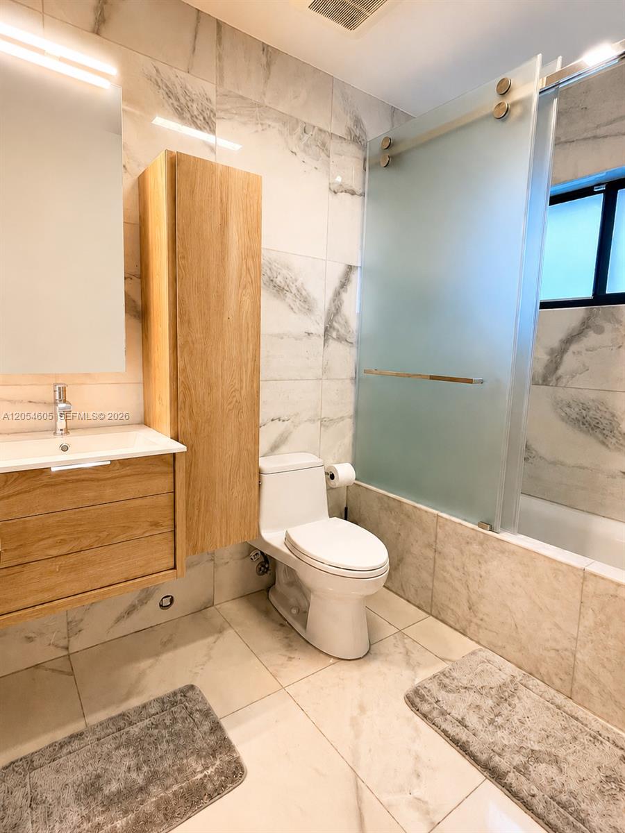 3538 Southwest 1st Avenue, Unit 1 Miami, FL 33145 - Photo 11 of 20 Second en-suite bathroom featuring marble-style finishes, a floating wood vanity with matching storage cabinet, illuminated mirror, modern toilet, and a glass-enclosed tub and shower combination with a frosted sliding door. A window provides natural light, while the neutral palette creates a clean, contemporary feel.