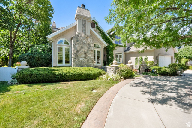 7740 Keswick Road Sandy, UT 84093 - Photo 4 of 74 Stucco exterior featuring a prominent stone chimney, arched windows, manicured landscaping, a concrete driveway with a decorative border, and a white picket fence