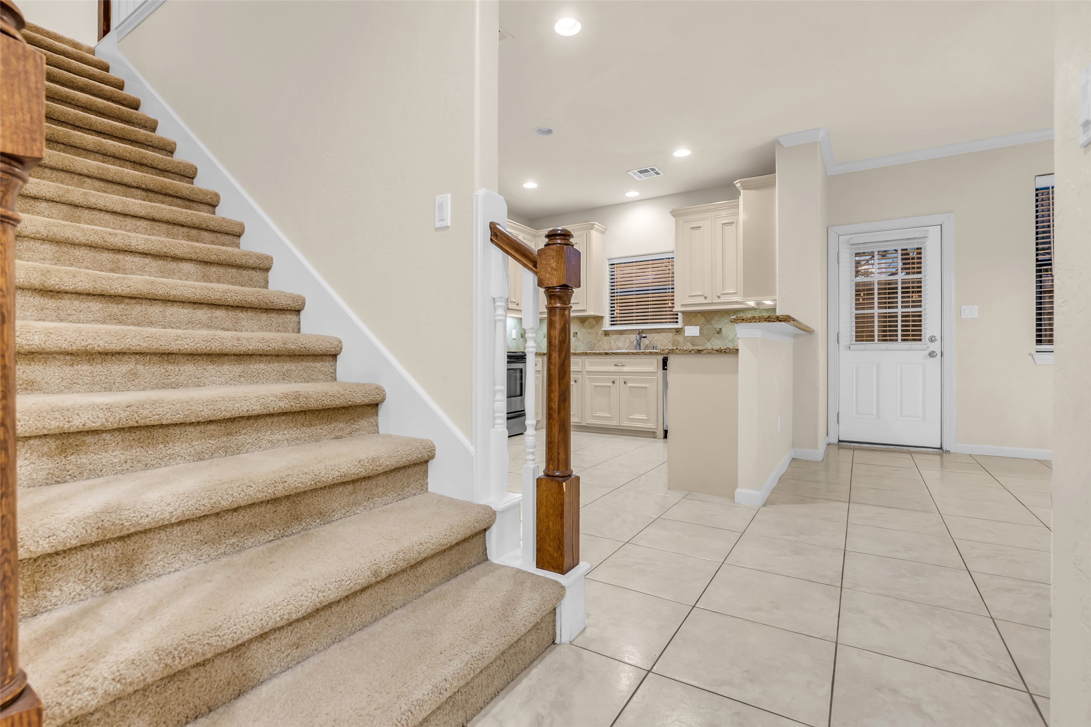 1898 Longmire Road, Unit 5 Conroe, TX 77304 - Photo 16 of 48 This photo showcases a welcoming entryway with carpeted stairs and a view into a bright, tiled kitchen area. The kitchen features light cabinetry, granite countertops, and access to an exterior door, offering a functional and inviting space.