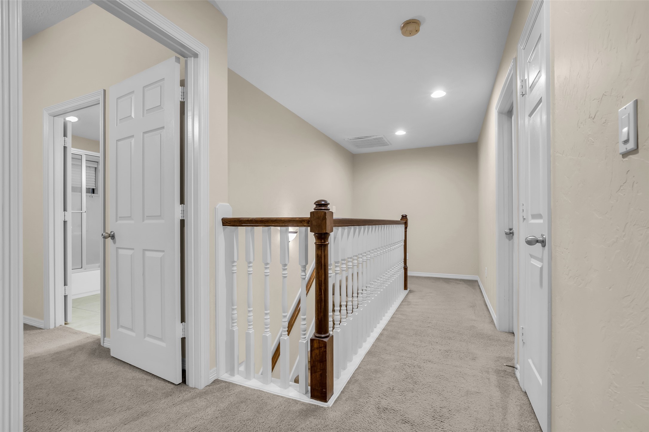 1898 Longmire Road, Unit 5 Conroe, TX 77304 - Photo 31 of 48 This photo shows a carpeted hallway with beige walls and recessed lighting. It features a wooden railing along a staircase and several white doors leading to other rooms, including a bathroom visible on the left.