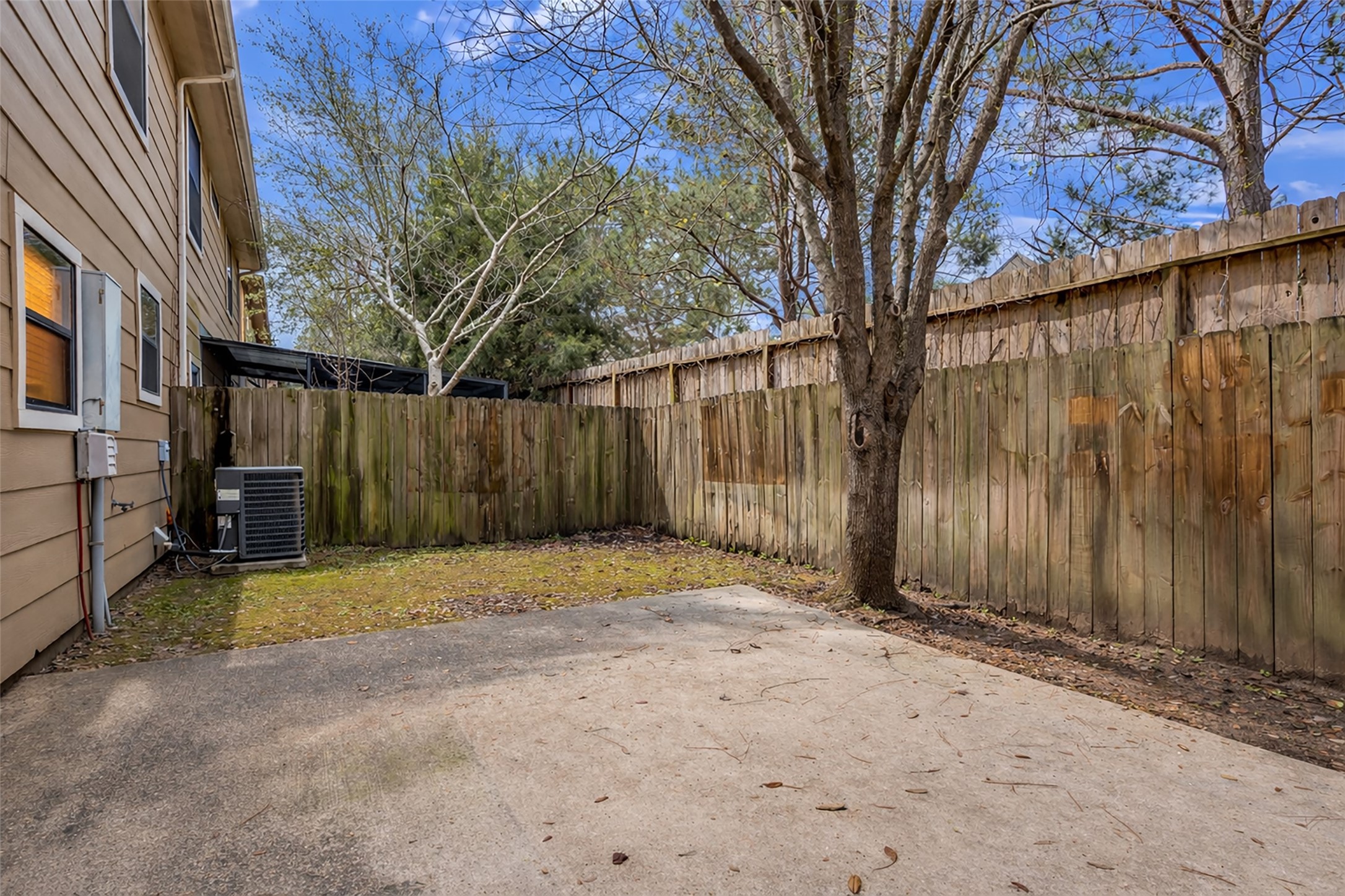 1898 Longmire Road, Unit 5 Conroe, TX 77304 - Photo 44 of 48 This photo shows a small, fenced backyard with a concrete patio and a tree, offering a private outdoor space for relaxation or entertaining. The area is adjacent to the house, with an air conditioning unit visible, and surrounded by wooden fencing.