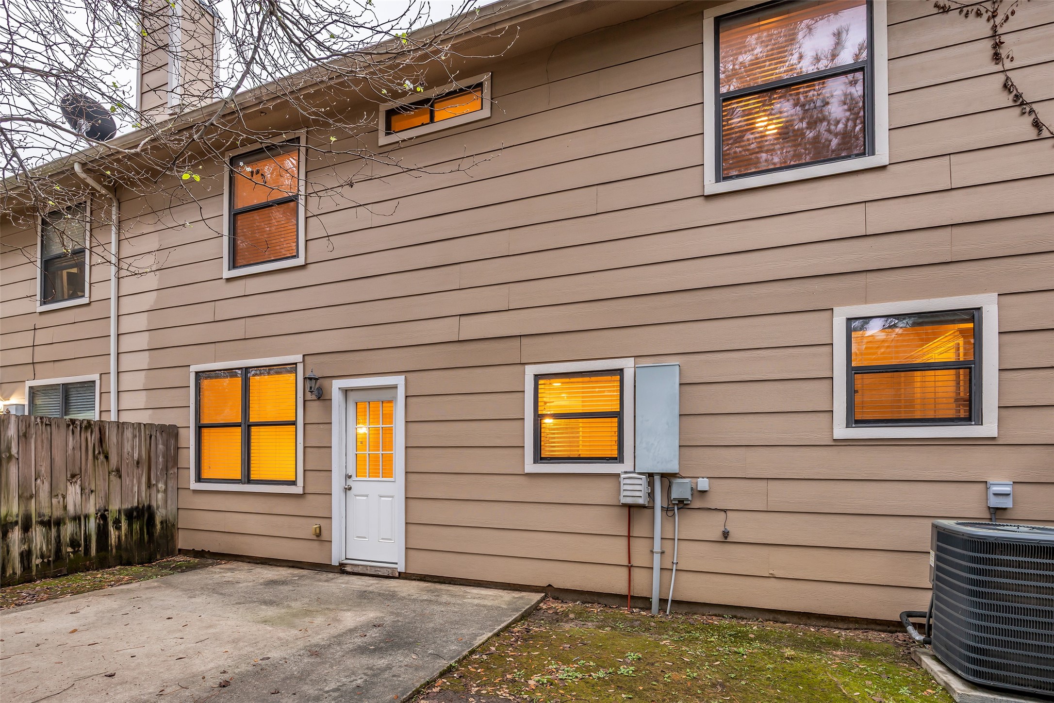 1898 Longmire Road, Unit 5 Conroe, TX 77304 - Photo 46 of 48 This photo shows the back exterior of a two-story home with tan siding and multiple windows. A small concrete patio area is adjacent to a wooden fence, and an air conditioning unit is visible. The area has some trees and natural surroundings.