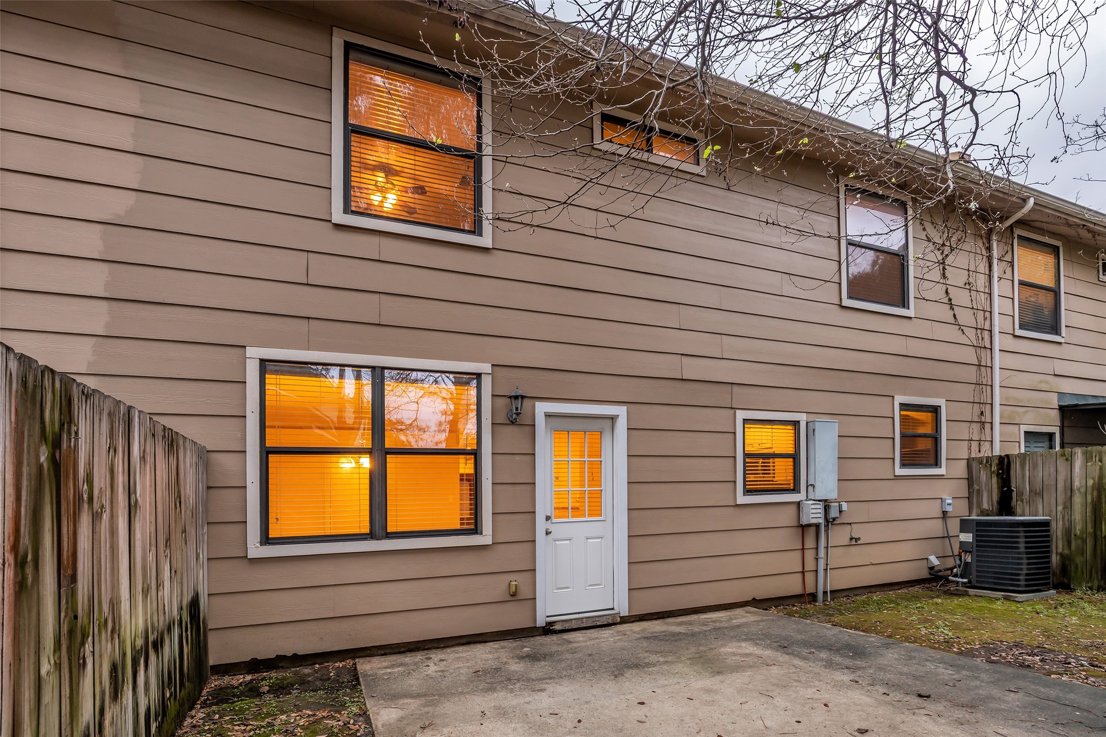 1898 Longmire Road, Unit 5 Conroe, TX 77304 - Photo 47 of 48 This photo shows the exterior of a two-story home with beige siding, multiple windows, and a back door. There's a small paved patio area and a wooden fence providing privacy. The windows have warm lighting from inside, and there's an air conditioning unit on the side.