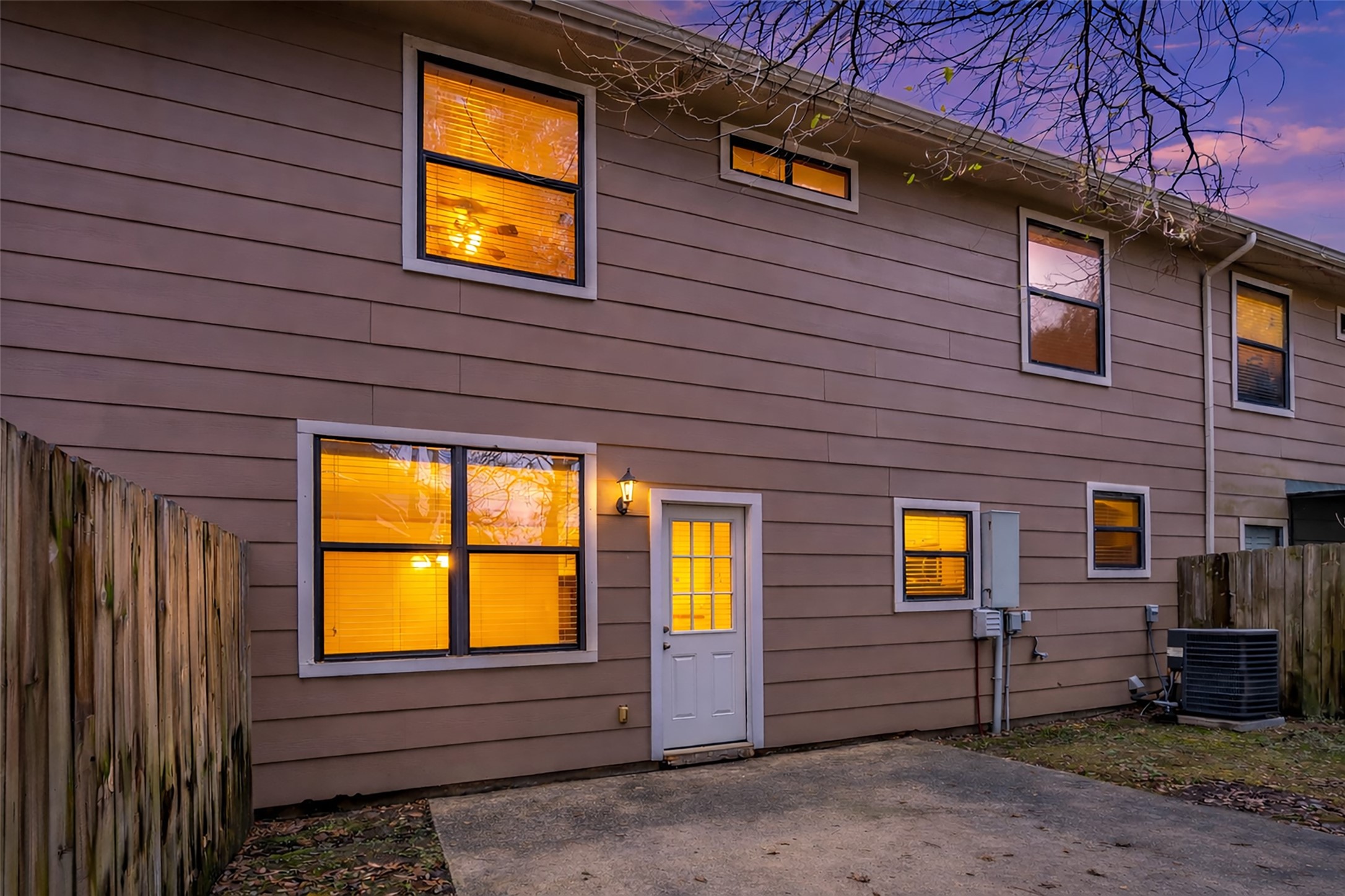 1898 Longmire Road, Unit 5 Conroe, TX 77304 - Photo 8 of 48 This photo shows the back of a two-story home at dusk, featuring a fenced yard, a concrete patio, and several windows warmly lit from inside. The siding is a neutral tone, and there is an exterior door leading to the backyard.