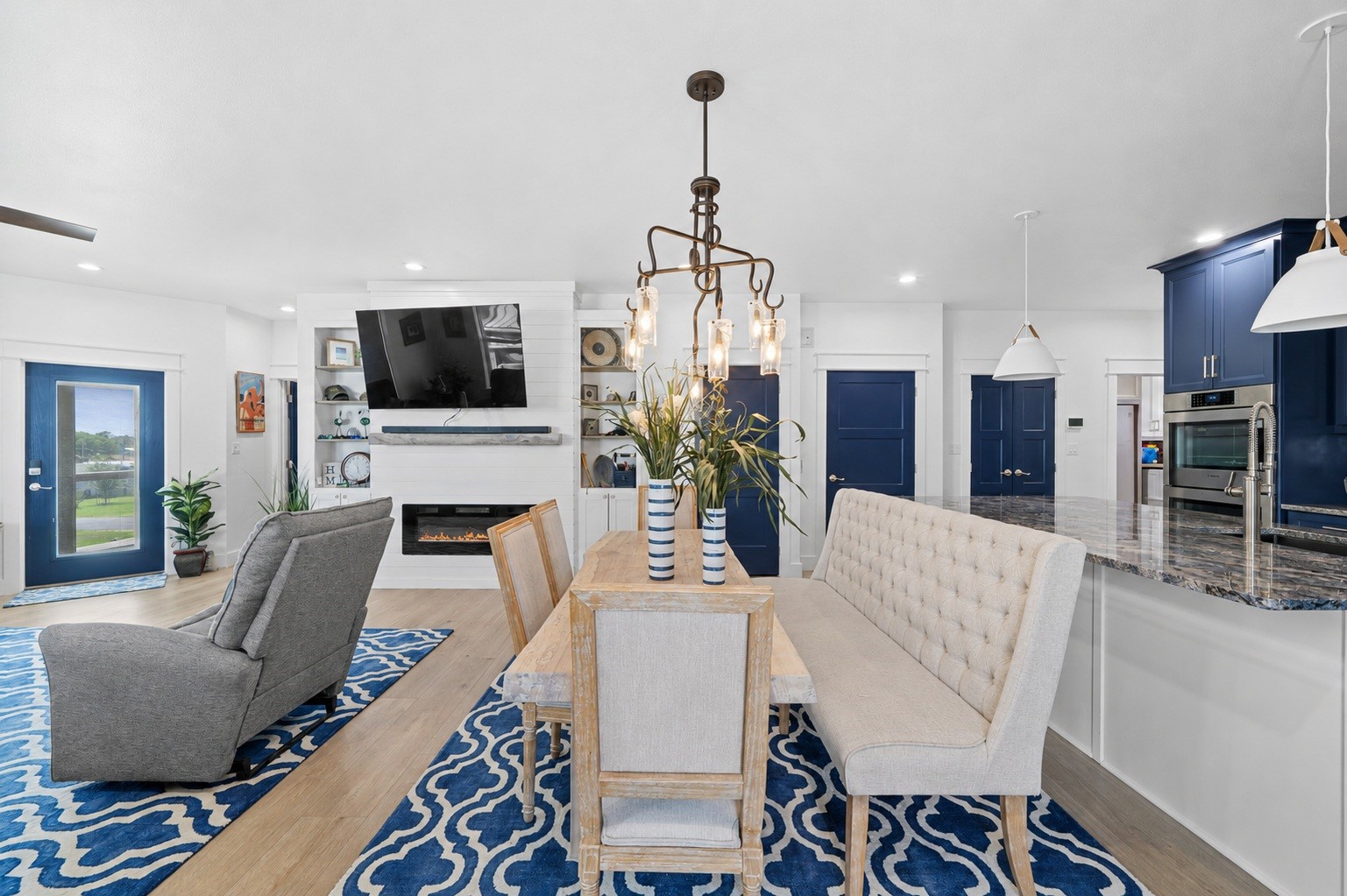29 Rabbit Drive Port O'Connor, TX 77982 - Photo 4 of 21 Spacious open-concept home interior featuring light hardwood floors, crisp white finishes, and deep navy blue accents. Highlights include a custom dining set with a tufted dining bench beneath an artistic chandelier.