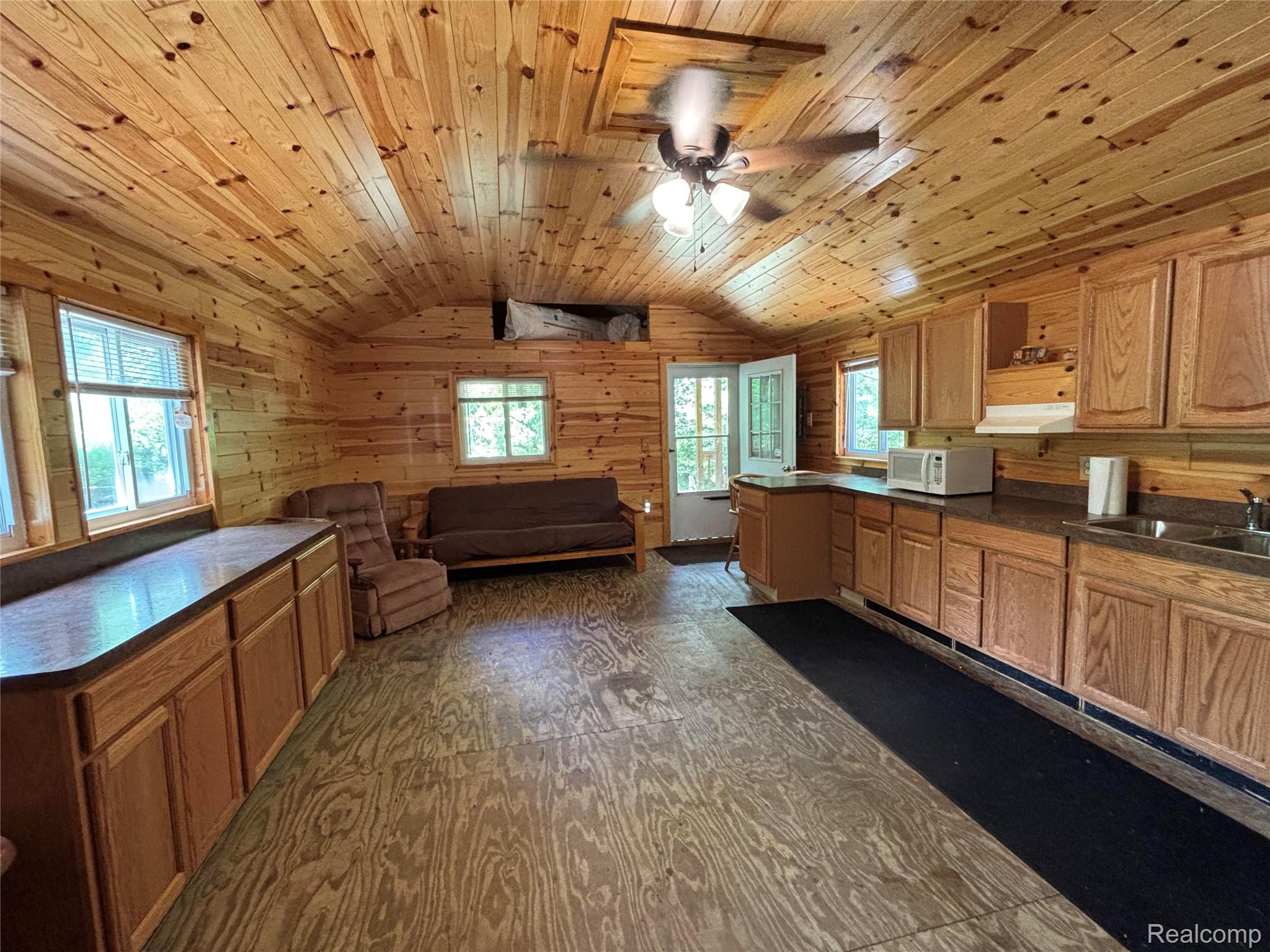 955 Aeronca Road Luzerne, MI 48636 - Photo 9 of 20 Open-concept living space featuring extensive wood-paneled walls and ceiling, a functional kitchen area with wood cabinetry and countertop space, a ceiling fan with integrated lighting, and multiple windows for natural light