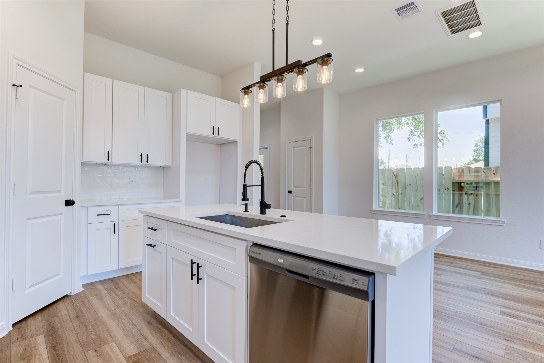 6809 Paris Street Houston, TX 77021 - Photo 12 of 37 Light-filled kitchen with a generous quartz island, single-basin sink, and matte black fixtures. Crisp white cabinetry and a subway tile backsplash keep it fresh, while pendant lighting adds warmth. A window brings in natural light, and the door to the left opens to a spacious pantry.