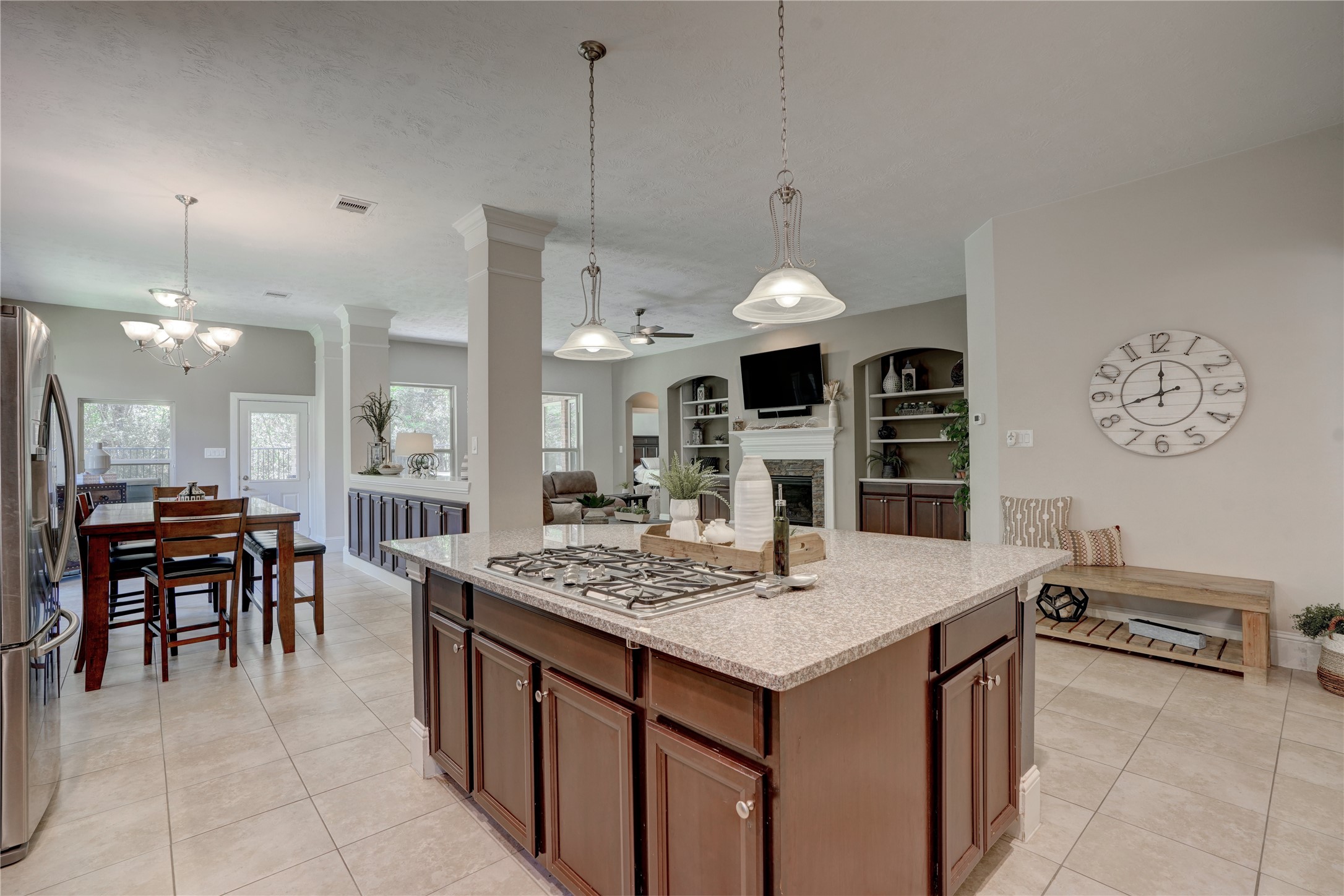 27243 Jessica Hills Lane Spring, TX 77386 - Photo 15 of 49 This spacious open-concept kitchen and living area features a large island with a cooktop, modern pendant lighting, and ample cabinetry. Adjacent to the kitchen is a cozy family room with built-in shelves and a fireplace, complemented by a dining area with natural light from large windows. The layout is perfect for entertaining and family gatherings.