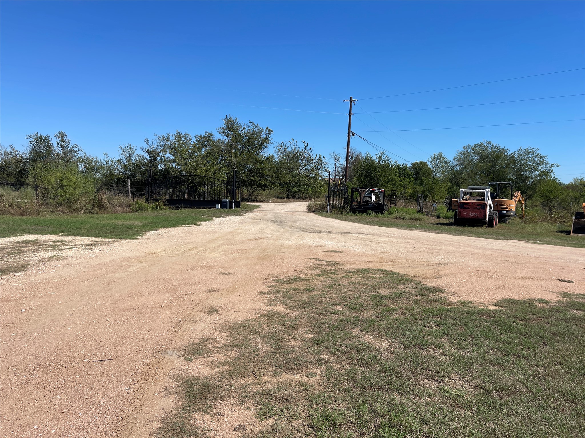 12212 Ballerstedt Road Elgin, TX 78621 - Photo 1 of 29 Gated access reveals a wide dirt driveway lined with mature trees under clear blue skies.