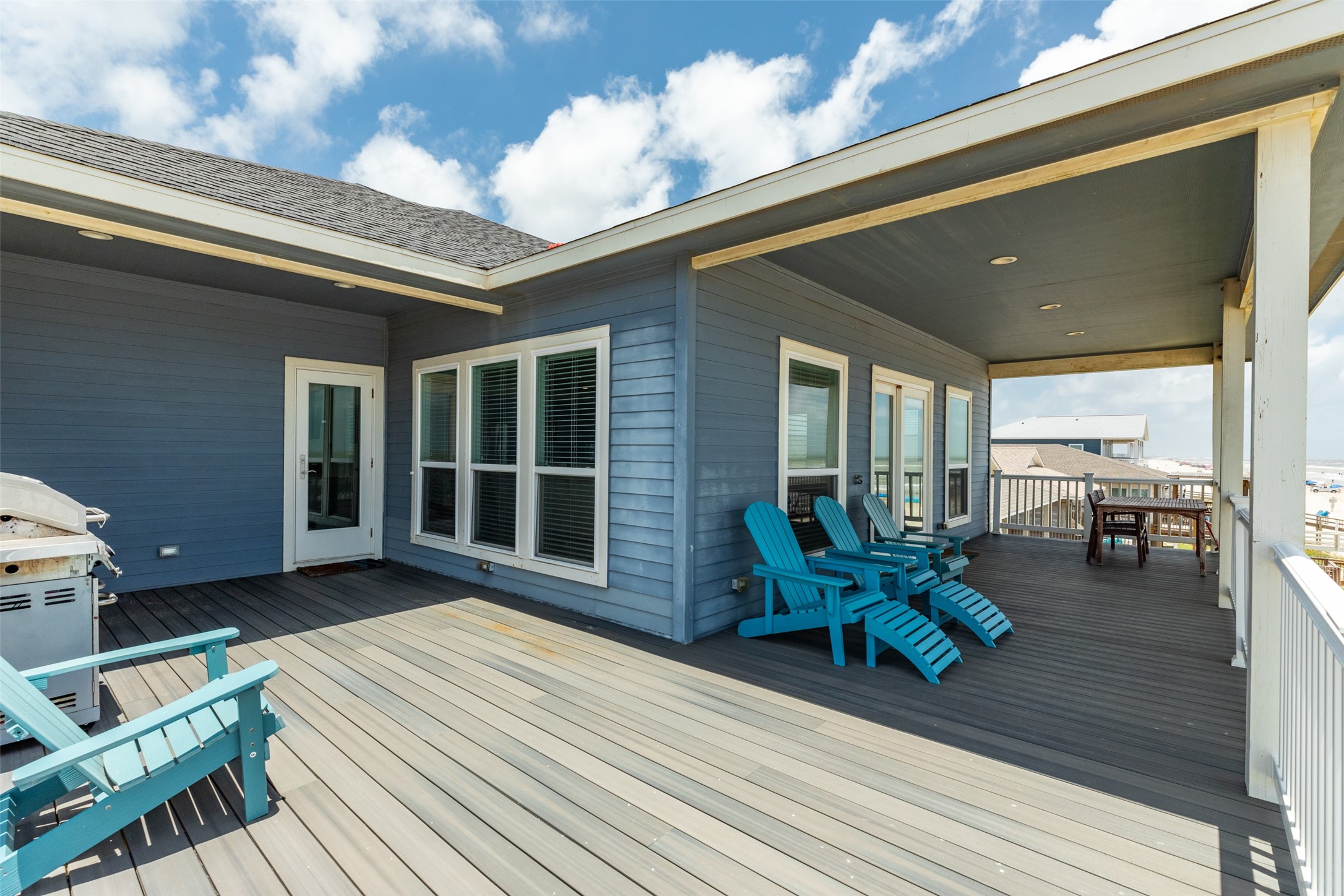 109 A Santar Loop, Unit A Surfside Beach, TX 77541 - Photo 18 of 43 This photo features a spacious, covered deck with blue siding and white trim, offering ample outdoor seating including Adirondack chairs. It includes a grill for outdoor cooking and provides a relaxed atmosphere with views of the surrounding area. Ideal for entertaining and enjoying sunny days.