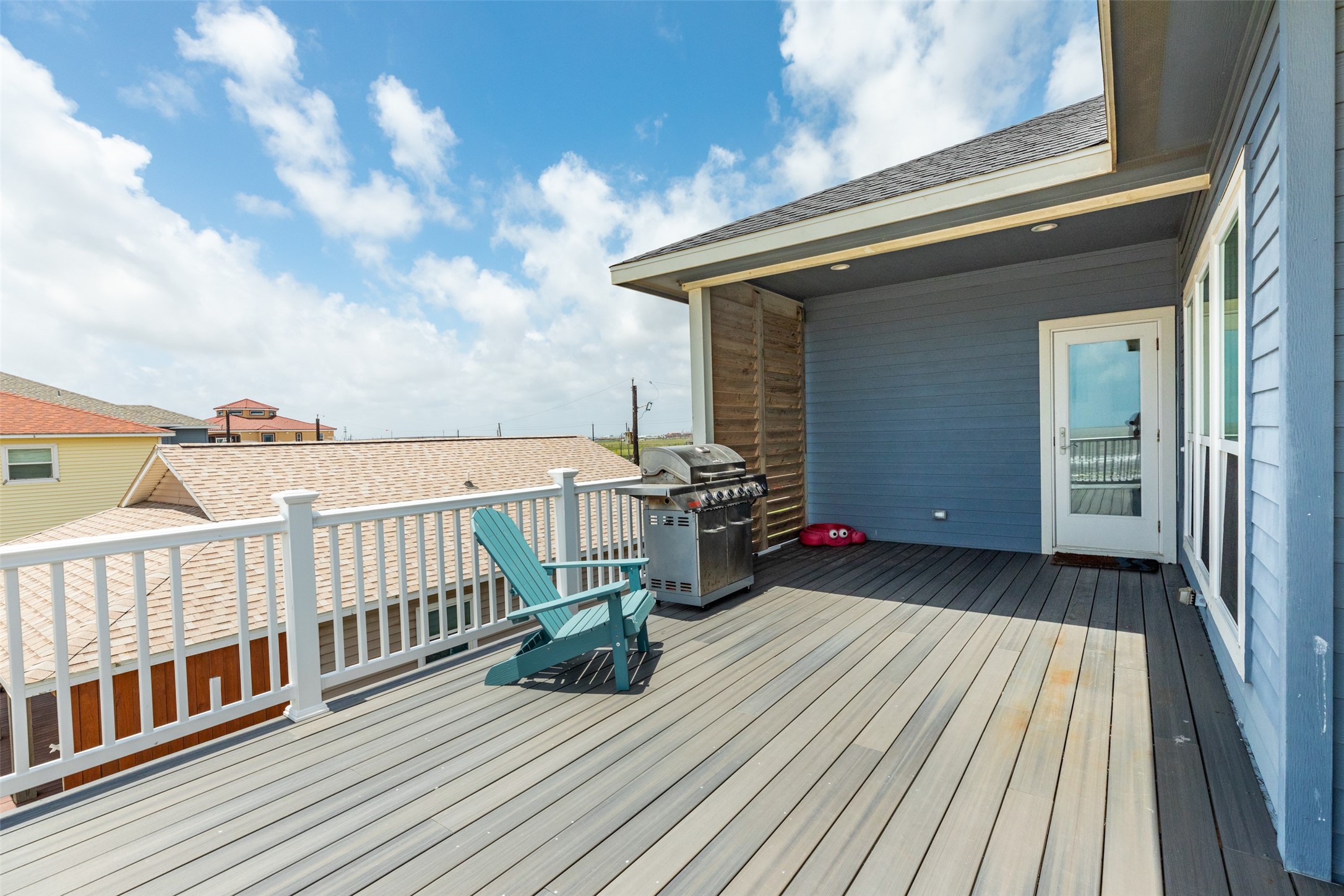 109 A Santar Loop, Unit A Surfside Beach, TX 77541 - Photo 19 of 43 This photo showcases a spacious, sunlit deck with modern gray flooring, a white railing, and a grill, perfect for outdoor entertaining. The deck offers views of neighboring rooftops under a bright blue sky.