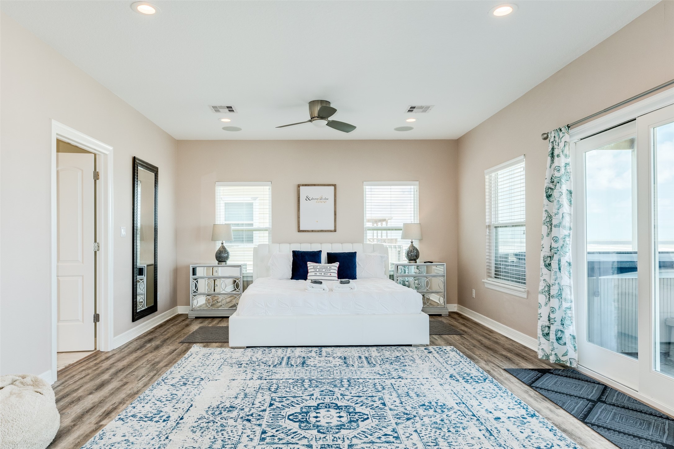 109 A Santar Loop, Unit A Surfside Beach, TX 77541 - Photo 24 of 43 This spacious bedroom features a modern design with neutral walls and wood-look flooring. It includes a large bed, mirrored nightstands, and ample natural light from multiple windows and glass doors leading to a deck. A ceiling fan and elegant rug complete the cozy atmosphere.