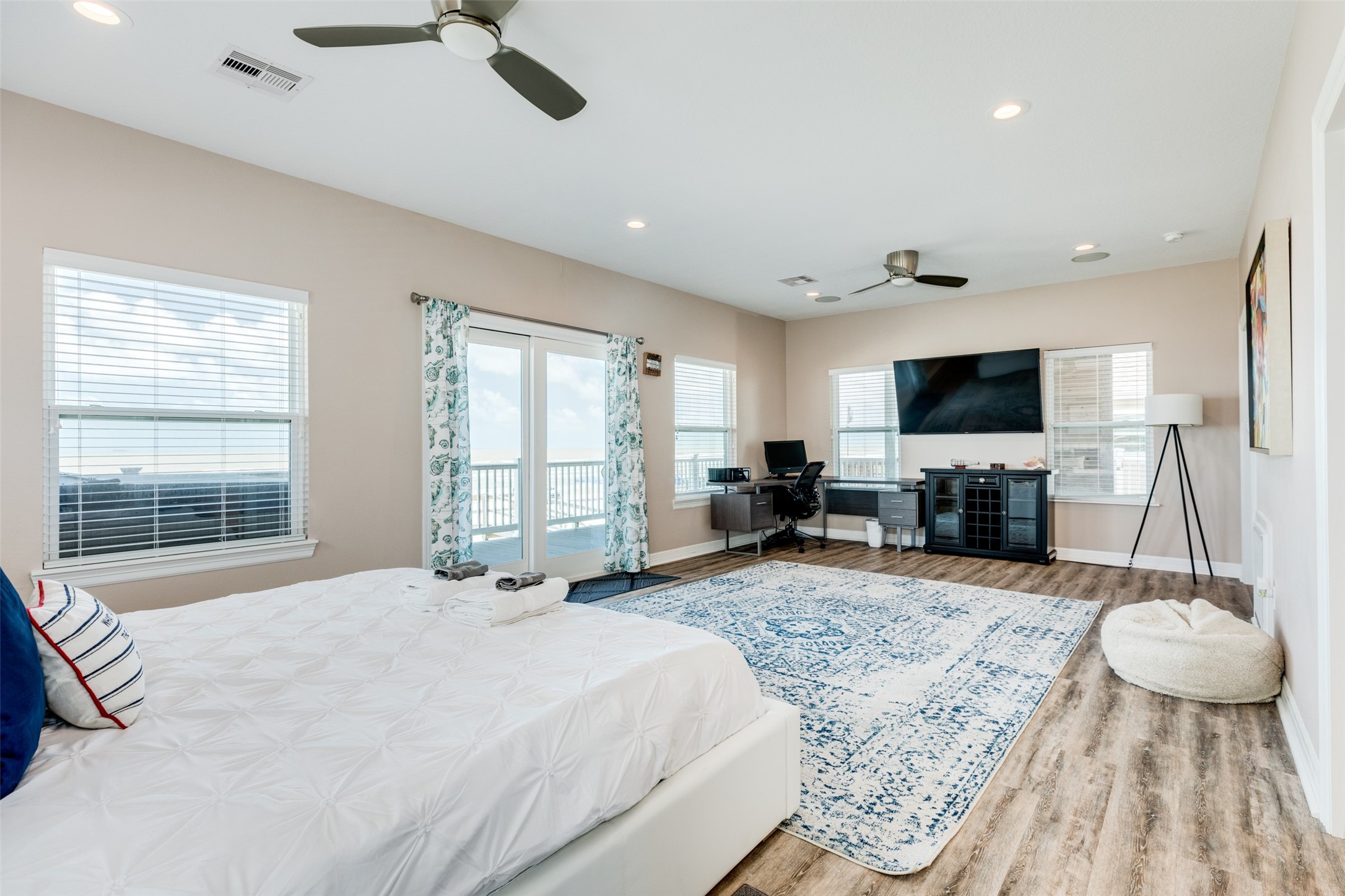 109 A Santar Loop, Unit A Surfside Beach, TX 77541 - Photo 25 of 43 Spacious, bright bedroom with large windows and glass doors leading to a deck. Features include wood flooring, modern ceiling fans, a cozy seating area, and a workspace with a large TV. Ideal for comfort and style.