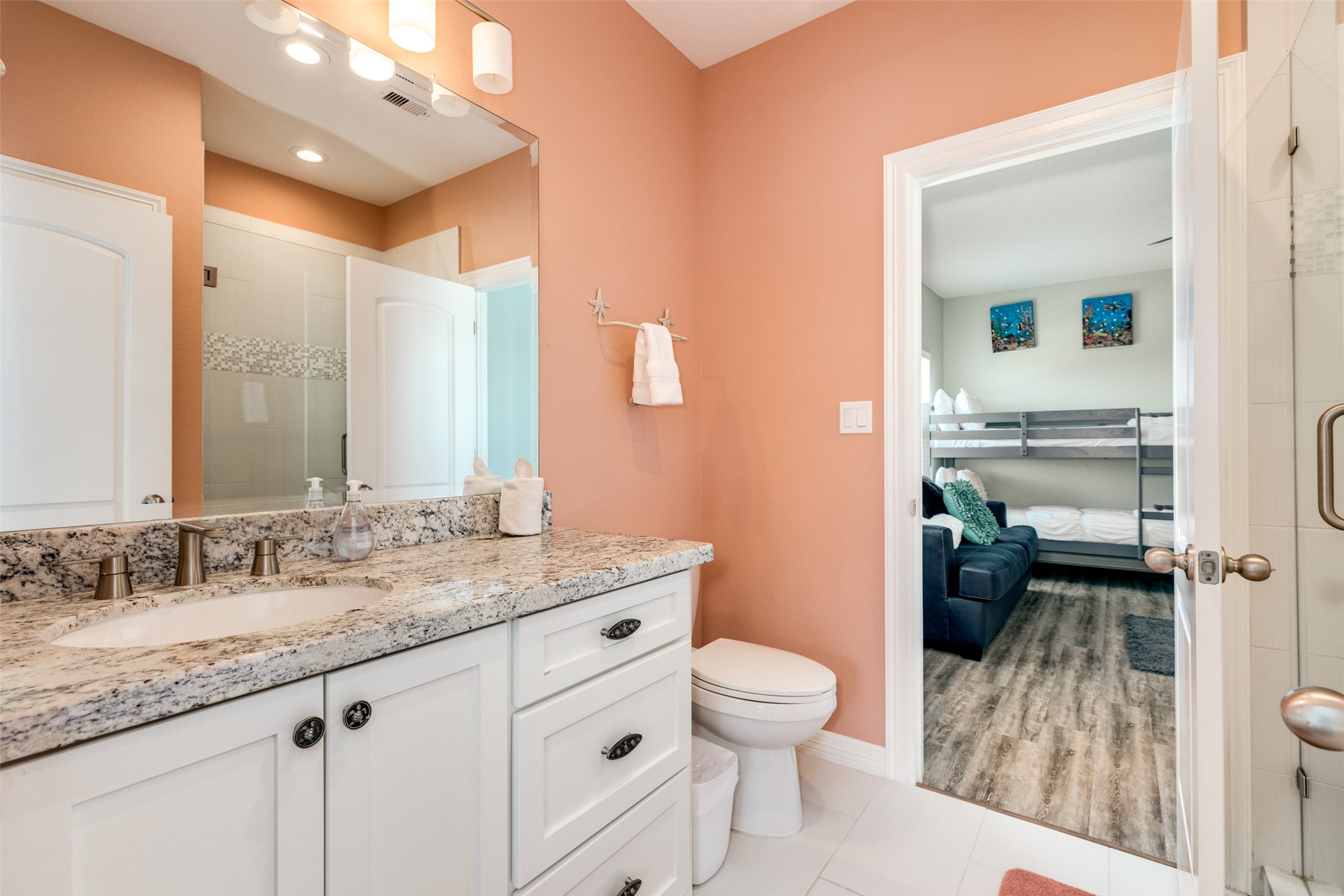 109 A Santar Loop, Unit A Surfside Beach, TX 77541 - Photo 30 of 43 This photo shows a modern bathroom with a granite countertop and dual sinks, leading into a bedroom with bunk beds and a cozy seating area. The walls are painted in warm tones, and the flooring is a combination of tile and wood look flooring.