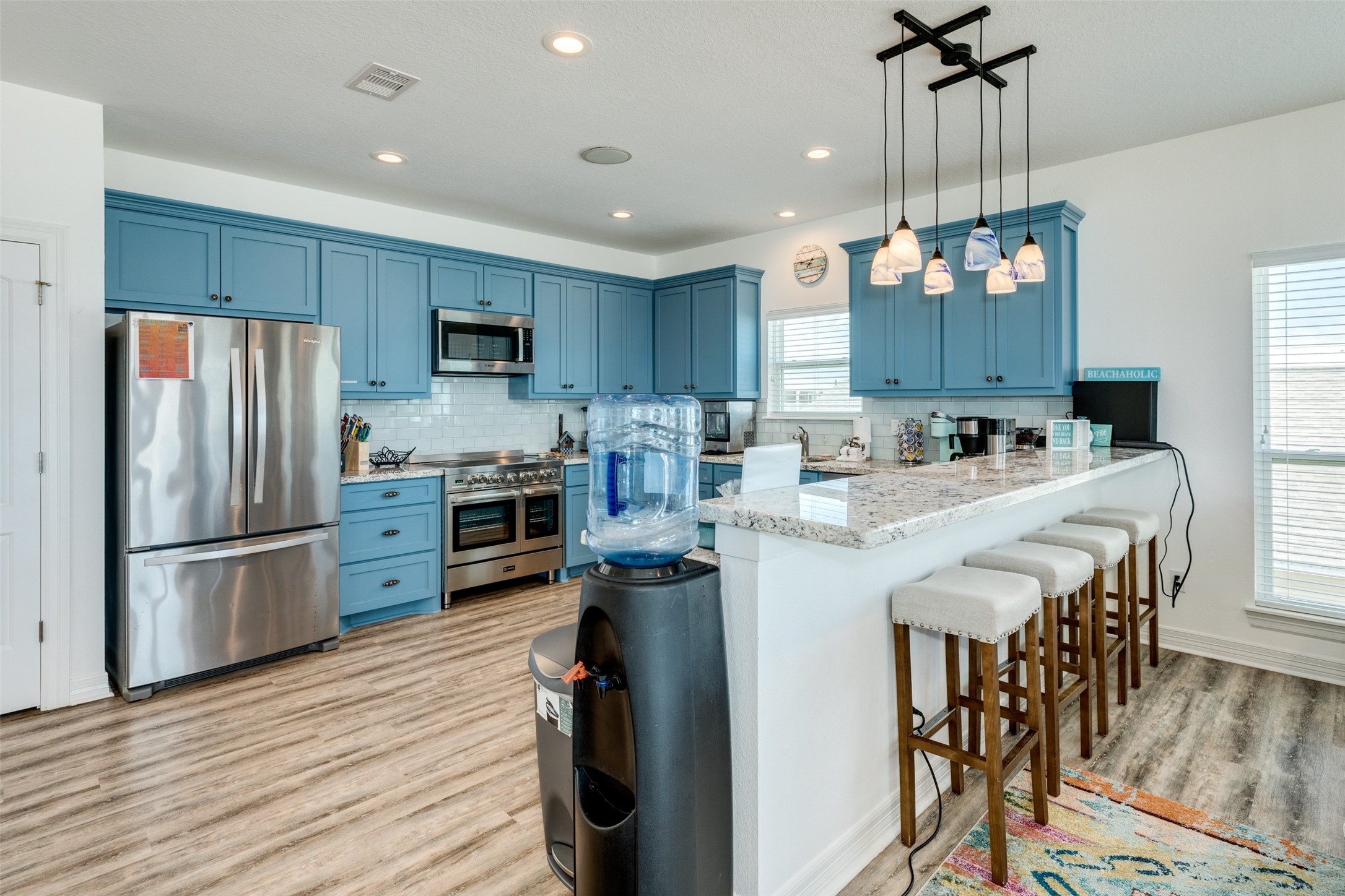 109 A Santar Loop, Unit A Surfside Beach, TX 77541 - Photo 34 of 43 This modern kitchen features vibrant blue cabinetry, stainless steel appliances, and a spacious countertop with bar seating. The room is well-lit with pendant lights and natural light from large windows, complemented by stylish flooring.