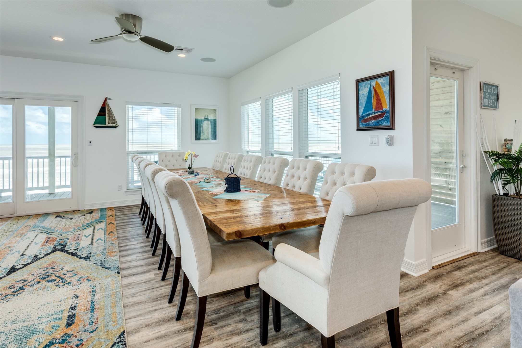 109 A Santar Loop, Unit A Surfside Beach, TX 77541 - Photo 36 of 43 Spacious, bright dining area with a long wooden table and cushioned chairs, large windows offering natural light, and a balcony view. Wood-style flooring and nautical decor create a coastal vibe.