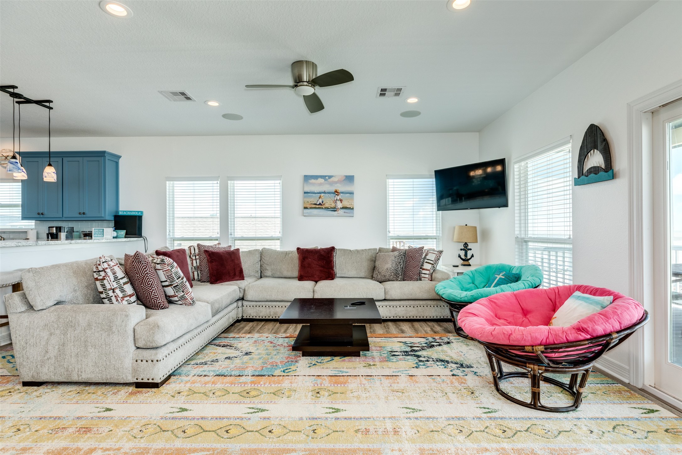 109 A Santar Loop, Unit A Surfside Beach, TX 77541 - Photo 38 of 43 Bright and airy living room with a large sectional sofa, colorful cushions, and a cozy atmosphere. Features a ceiling fan, modern decor, and ample natural light from multiple windows. Open view to the kitchen with blue cabinets.