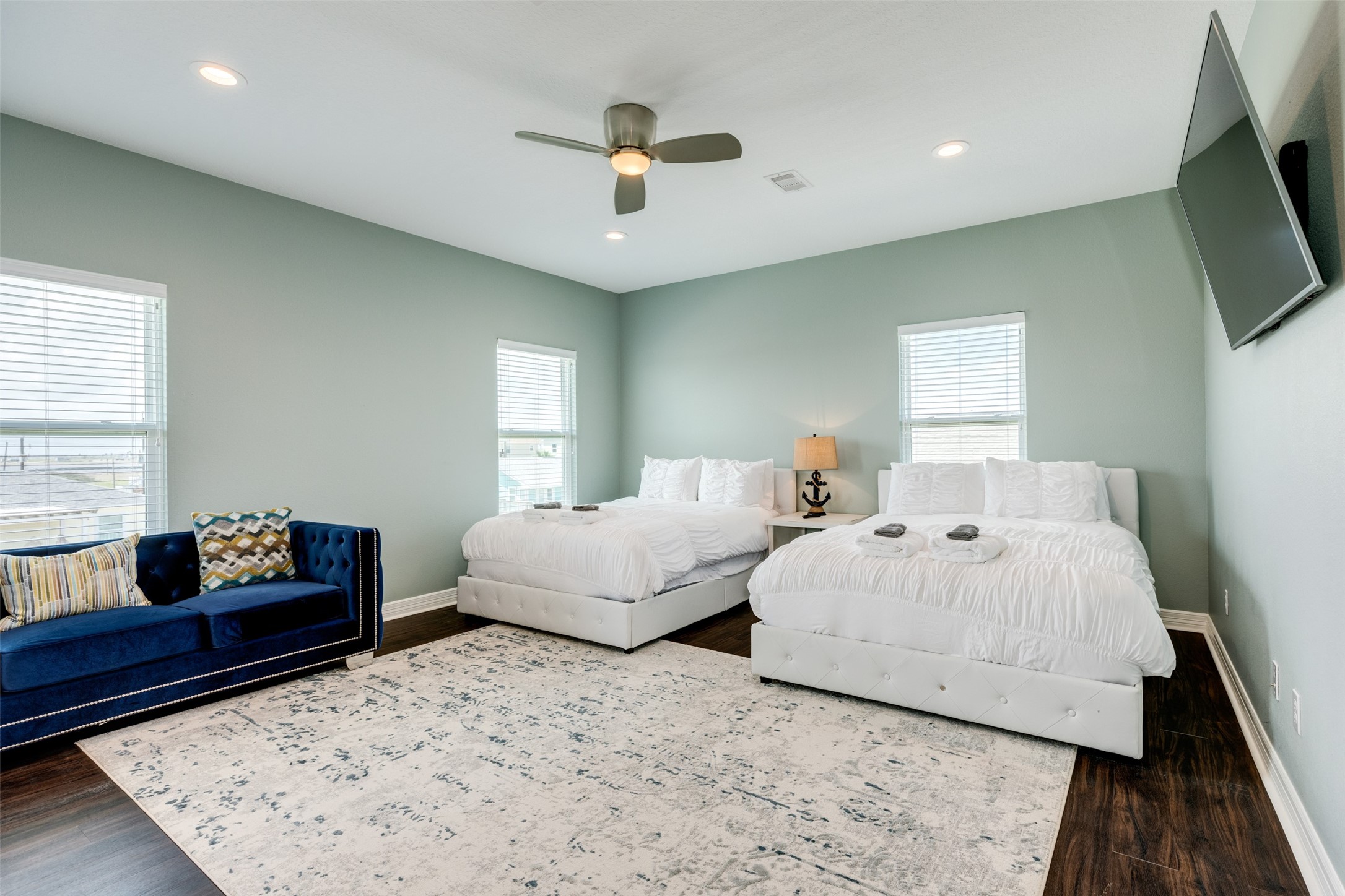 109 A Santar Loop, Unit A Surfside Beach, TX 77541 - Photo 39 of 43 Spacious bedroom with two beds, a cozy blue sofa, and a large TV. The room features soft green walls, dark hardwood floors, and ample natural light from two windows.