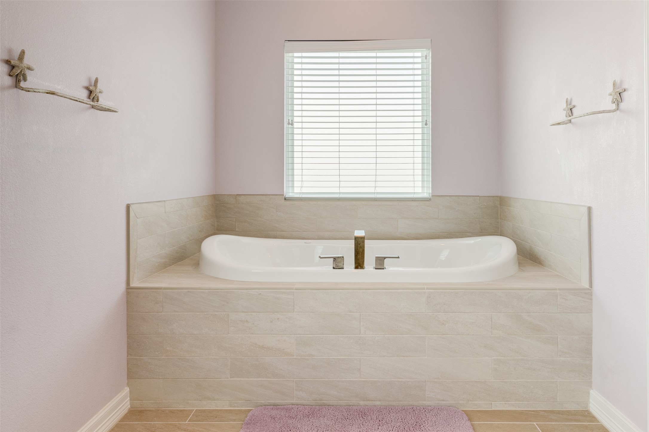 109 A Santar Loop, Unit A Surfside Beach, TX 77541 - Photo 42 of 43 This bathroom features a modern, built-in corner bathtub with a tiled surround, a window with blinds for natural light, and subtle lavender walls for a calming atmosphere.