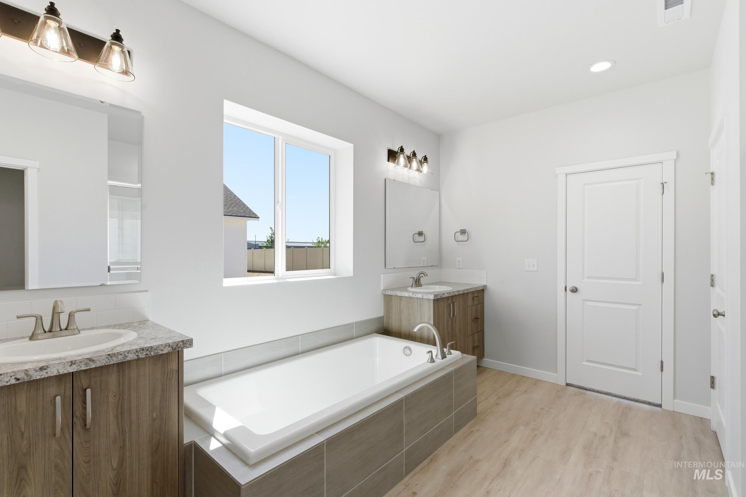 13663 Redway Court Caldwell, ID 83607 - Photo 31 of 32 Contemporary bathroom featuring dual wood-finish vanities with light stone-look countertops, a built-in soaking tub with tiled surround, a large window, and light wood-finish flooring