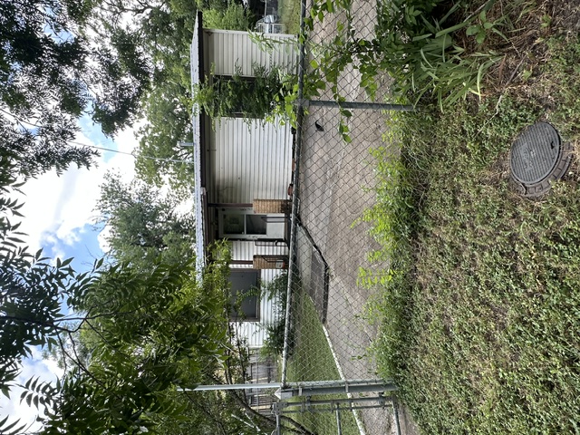 1127 Mark Street, Unit D Austin, TX 78721 - Photo 2 of 6 A modest structure with siding and a tin roof stands amidst lush greenery and a chain link fence.