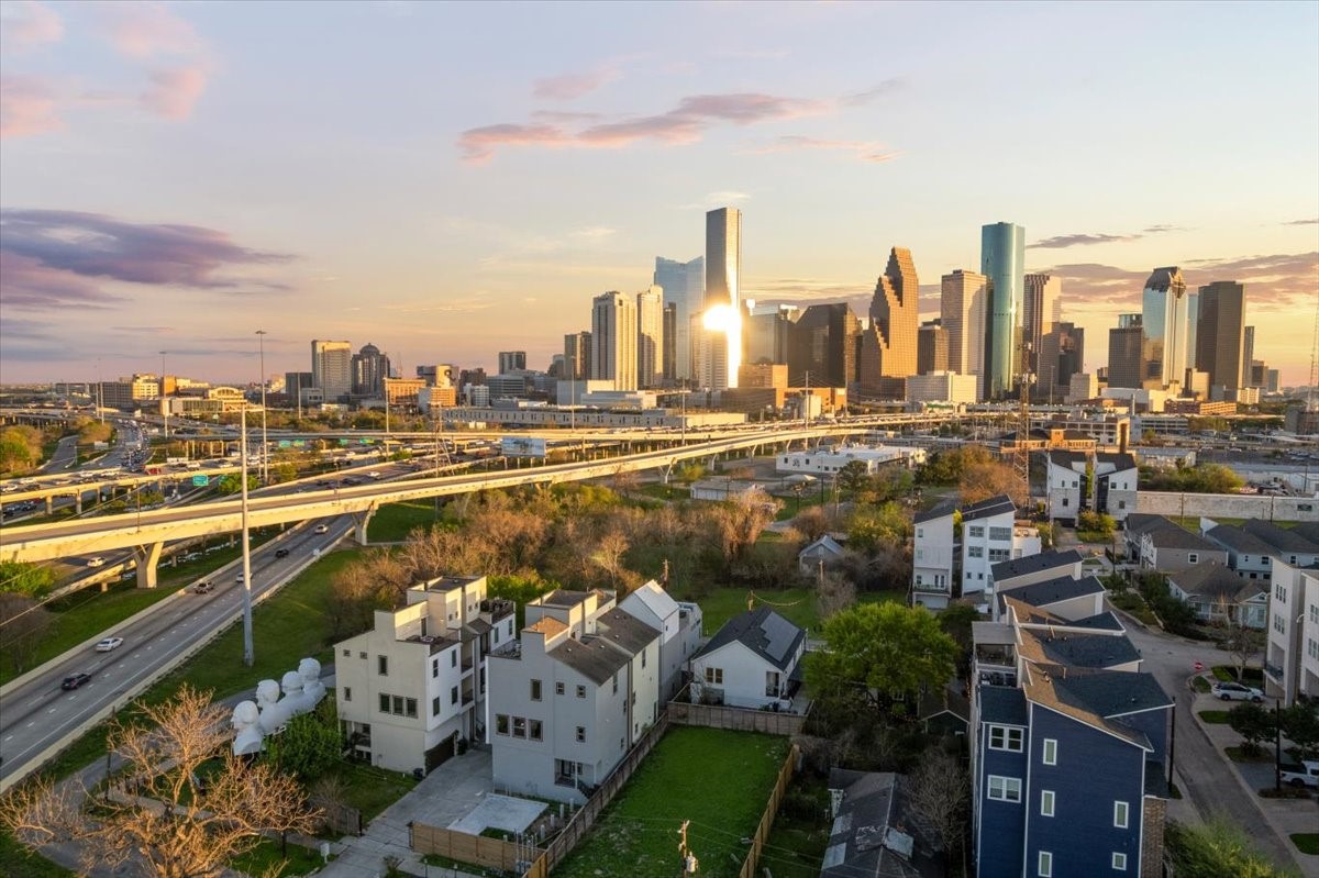 1005 Bingham Street, Unit 5 Houston, TX 77007 - Photo 14 of 50 This photo showcases a vibrant urban skyline with modern high-rises under a golden sunset. In the foreground, there are contemporary residential upscale homes surrounded by greenery, with easy access to major highways, suggesting a convenient location for city living.