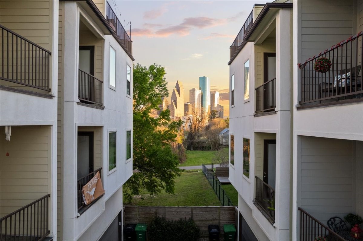 1005 Bingham Street, Unit 5 Houston, TX 77007 - Photo 17 of 50 This photo captures a view from a residential area with modern townhouses on either side, leading to a scenic view of a city skyline in the background. The setting includes a grassy area and trees, offering a blend of urban and natural surroundings.