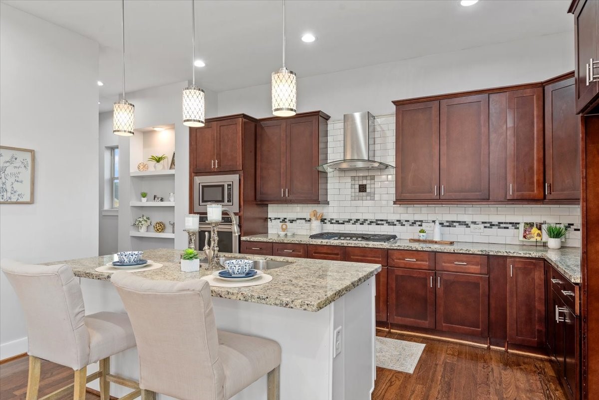 1005 Bingham Street, Unit 5 Houston, TX 77007 - Photo 2 of 50 This modern kitchen showcases a distinctive touch with decorative white shelving, beautifully illuminated from above to highlight special displays. Rich dark wood cabinets pairs elegantly with granite countertops, and a stylish subway tile backsplash. It includes a built-in microwave, stainless steel range hood, and a nice island with pendant lighting, creating a warm and inviting space perfect for cooking and entertaining.