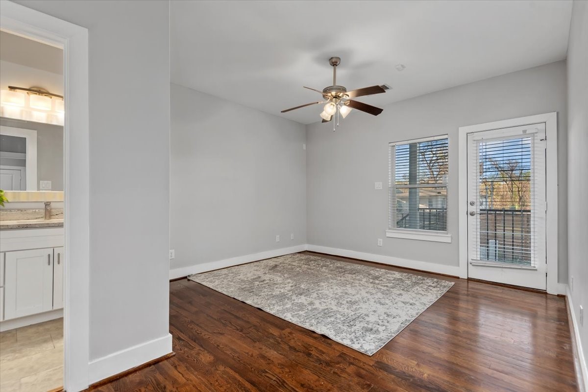 1005 Bingham Street, Unit 5 Houston, TX 77007 - Photo 25 of 50 This room features hardwood floors, neutral gray walls, and a ceiling fan with lights. There's a large window and a door with glass panels leading to a small outdoor space. A glimpse of a modern bathroom with white cabinetry is visible from the room.