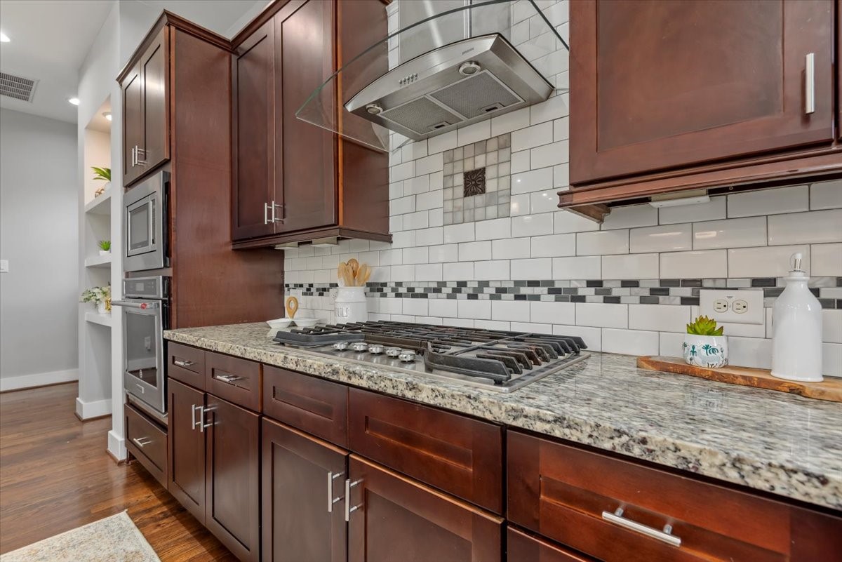 1005 Bingham Street, Unit 5 Houston, TX 77007 - Photo 32 of 50 This kitchen features elegant dark wood cabinetry, a modern gas stove with a sleek hood, granite countertops, and a stylish white subway tile backsplash with decorative accents. Perfect for culinary enthusiasts!