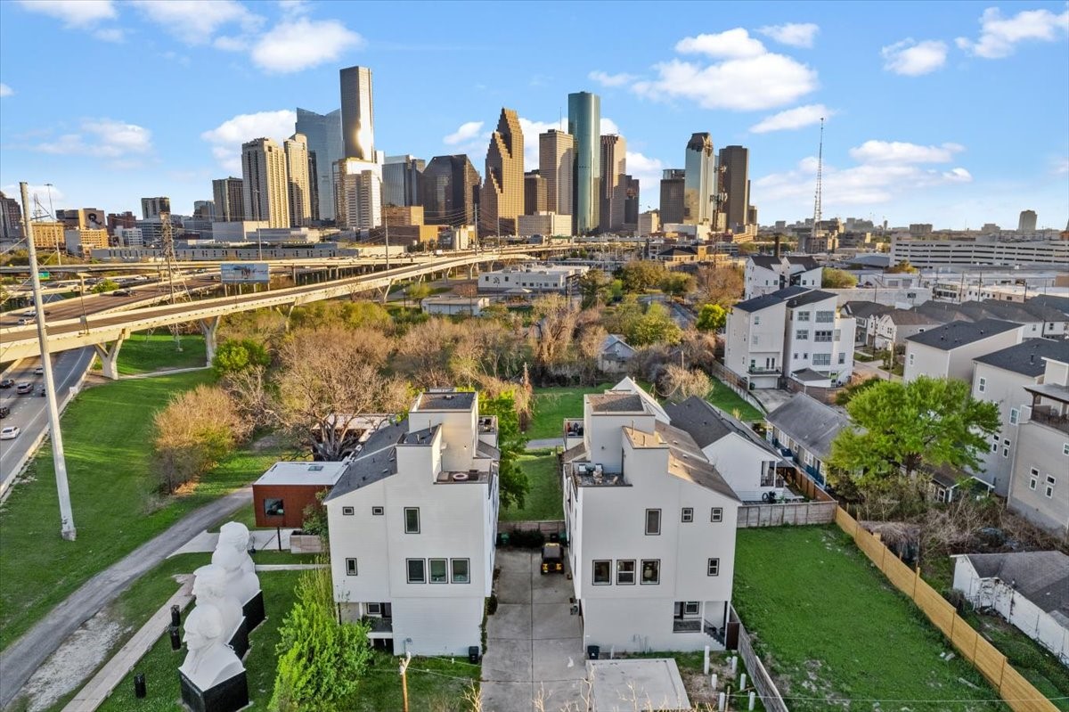 1005 Bingham Street, Unit 5 Houston, TX 77007 - Photo 44 of 50 This photo showcases a modern residential area with two white multi-story homes, set against a backdrop of a vibrant city skyline. The homes are surrounded by green spaces and are conveniently located near major roadways (I-10 and I-45), offering both urban and suburban living benefits.
