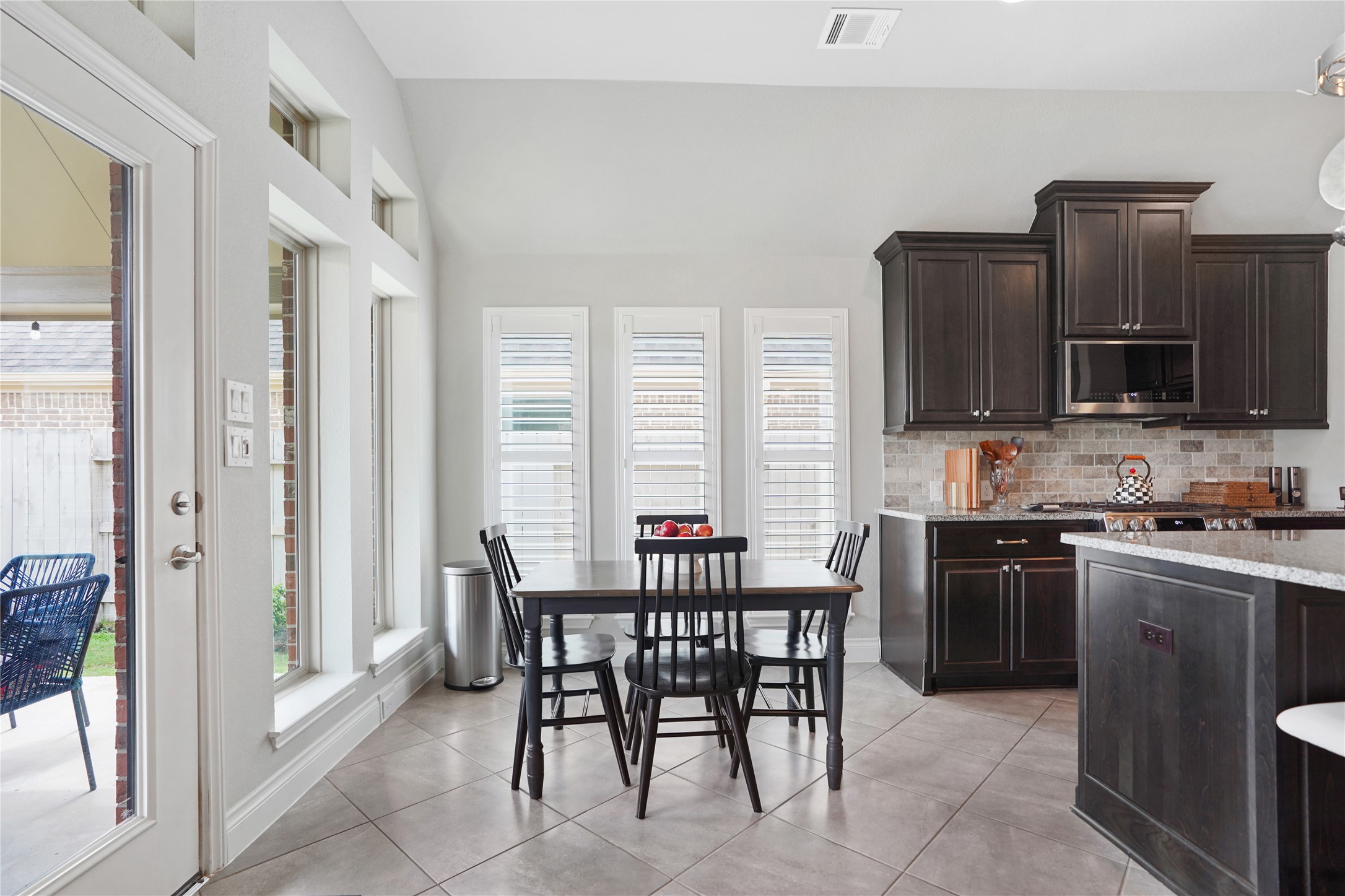 2345 Redwood Ridge Trail Manvel, TX 77578 - Photo 21 of 39 This photo showcases a bright kitchen with dark wooden cabinets, a cozy dining area, and large windows with white shutters. The space opens to a patio, providing an airy feel and outdoor access.