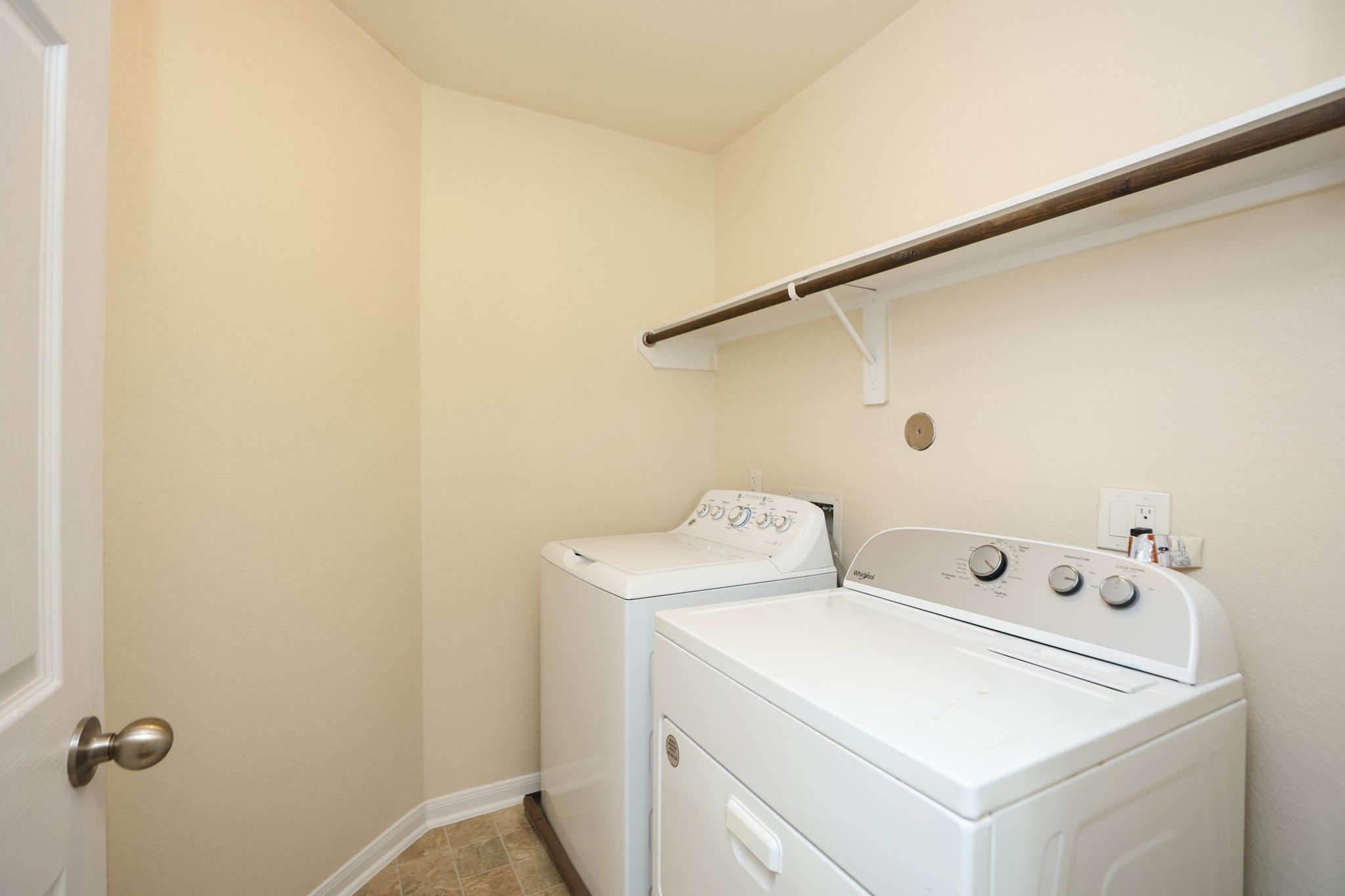 18402 Spruce Tree Line Trail Houston, TX 77084 - Photo 15 of 30 This photo shows a compact laundry room with a washer and dryer. It has a beige wall, tile flooring, and a wooden shelf above the appliances for storage.