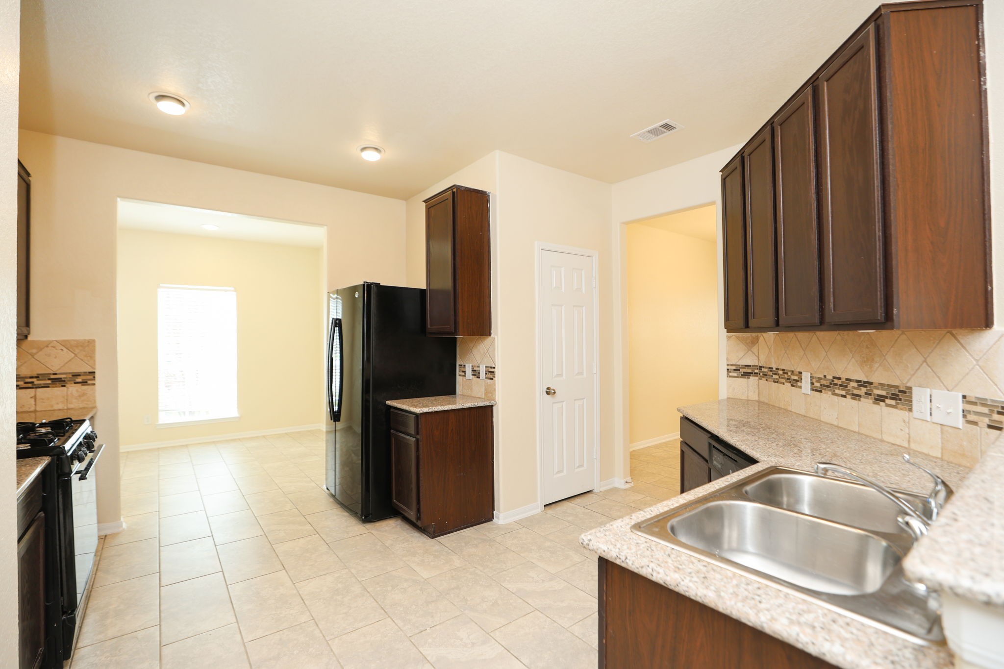 18402 Spruce Tree Line Trail Houston, TX 77084 - Photo 6 of 30 This kitchen features dark wood cabinets, a modern tile backsplash, and granite countertops. It includes a double sink, stainless steel appliances, and tiled flooring. A doorway leads to a bright dining area with natural light.