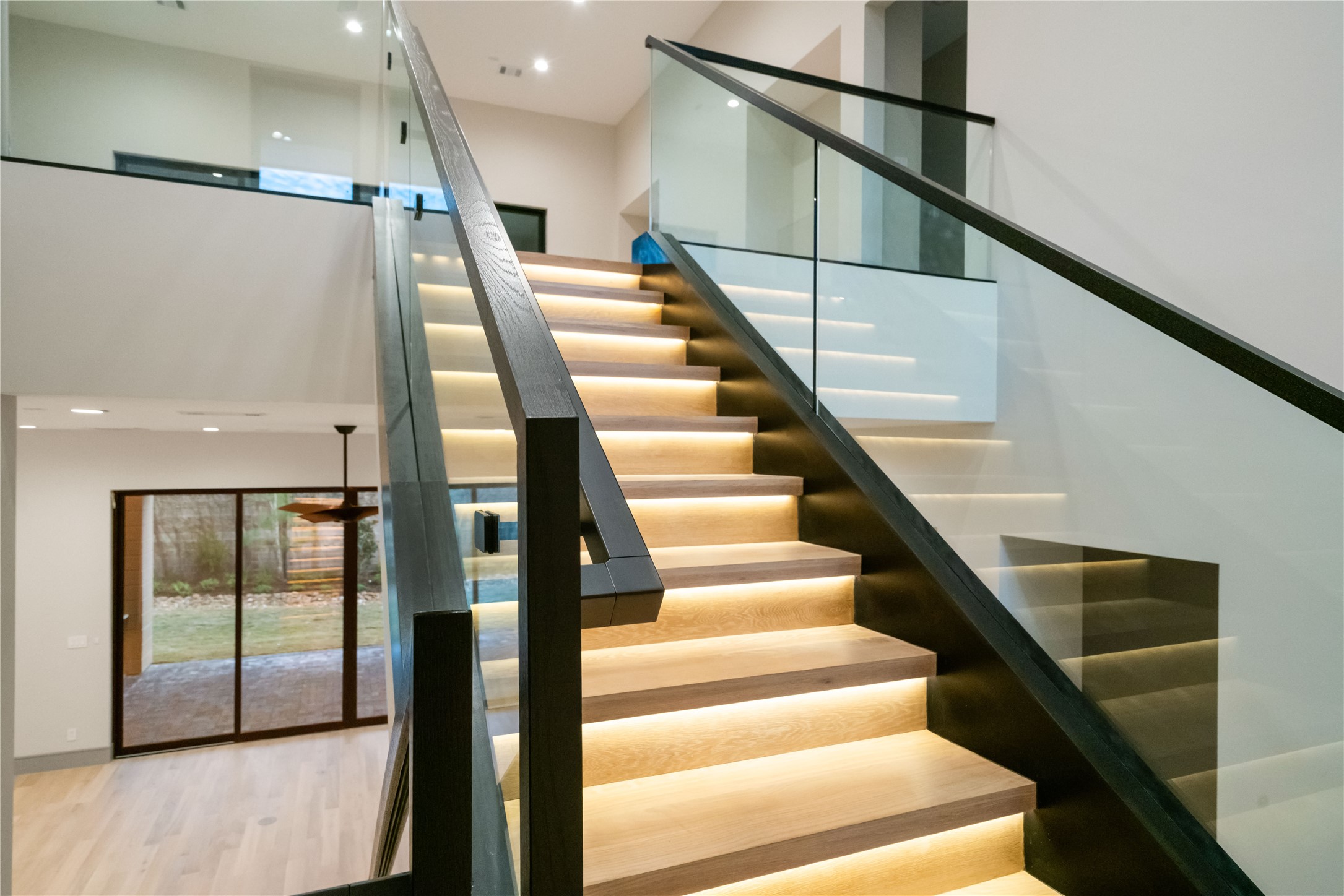47 Pronghorn Place Spring, TX 77389 - Photo 24 of 44 This photo showcases a modern staircase with sleek glass railings and elegant under-step lighting. The open layout includes a view of a spacious room with sliding glass doors leading to an outdoor space, highlighting natural light and contemporary design.