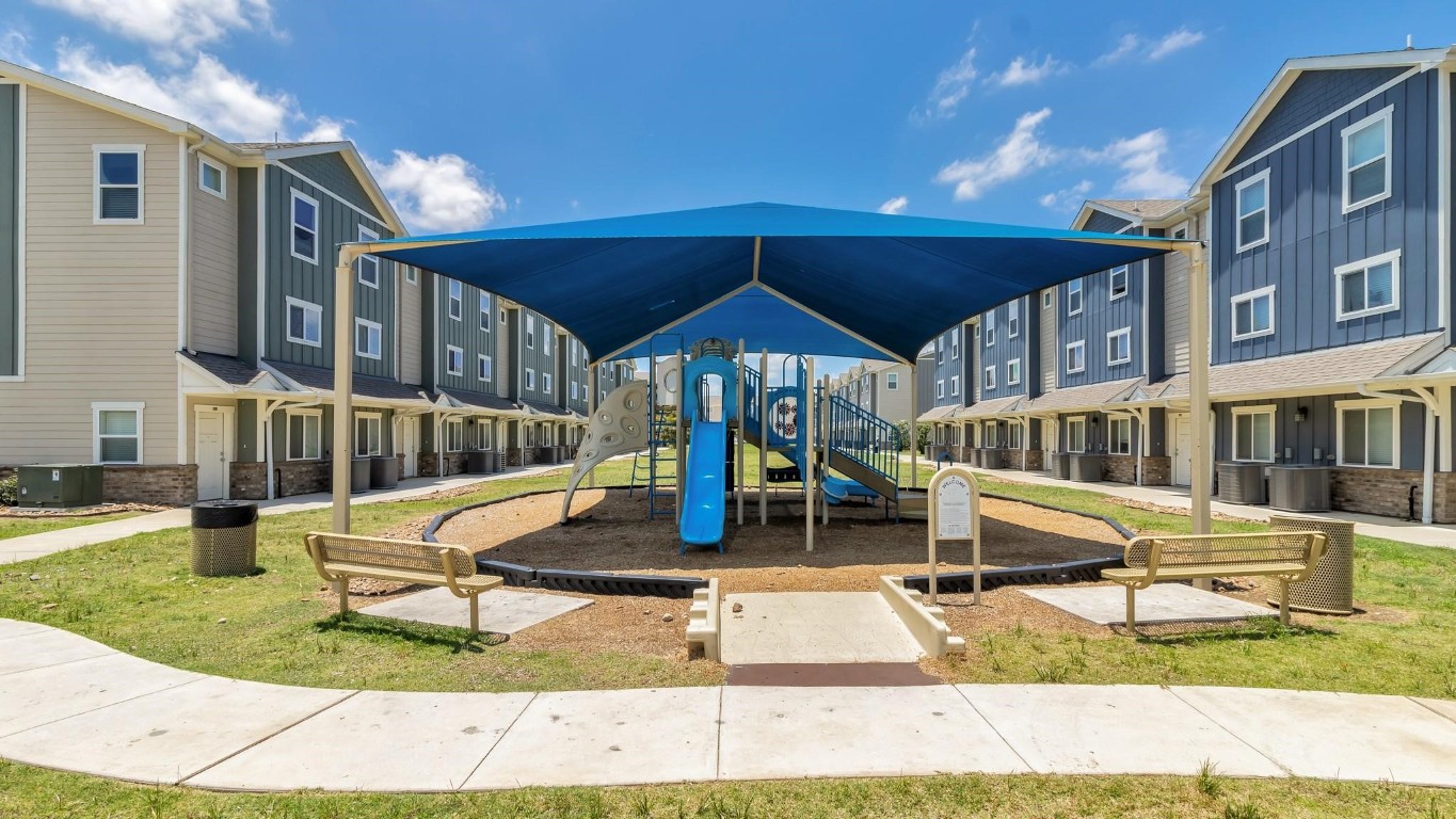21155 Gosling Road, Unit 15 AB Spring, TX 77388 - Photo 24 of 30 This photo shows a modern playground with a climbing structure and slides, sheltered by a blue canopy. It's set in a well-maintained grassy area with nearby residential buildings, ideal for families.