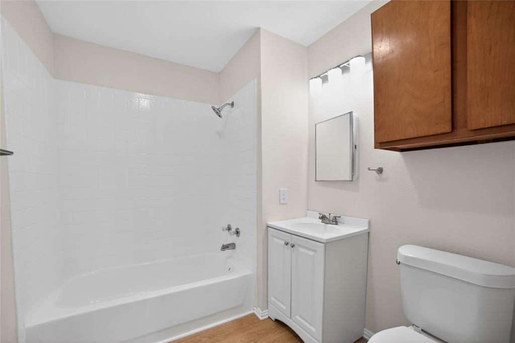 808 Park Street Commerce, TX 75428 - Photo 19 of 29 This bathroom features a shower and bathtub combination with white tiled walls, a white vanity with a sink, a wall-mounted mirror, and a toilet
