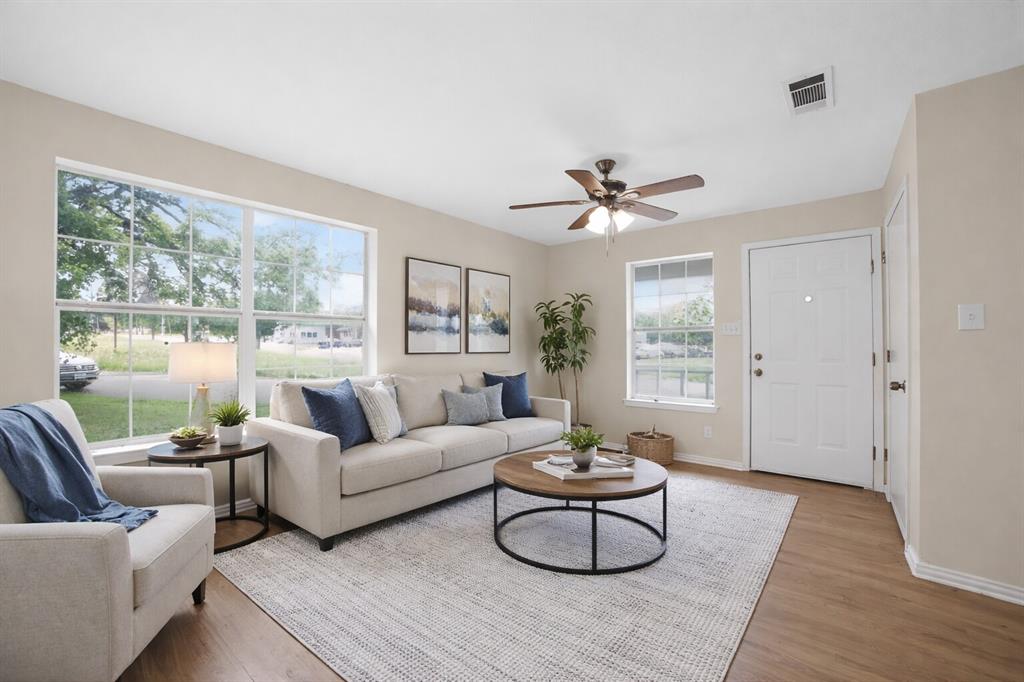 808 Park Street Commerce, TX 75428 - Photo 2 of 29 The living room features large windows, a ceiling fan with lighting, wood-style flooring, and a light-colored entry door - Virtually Staged