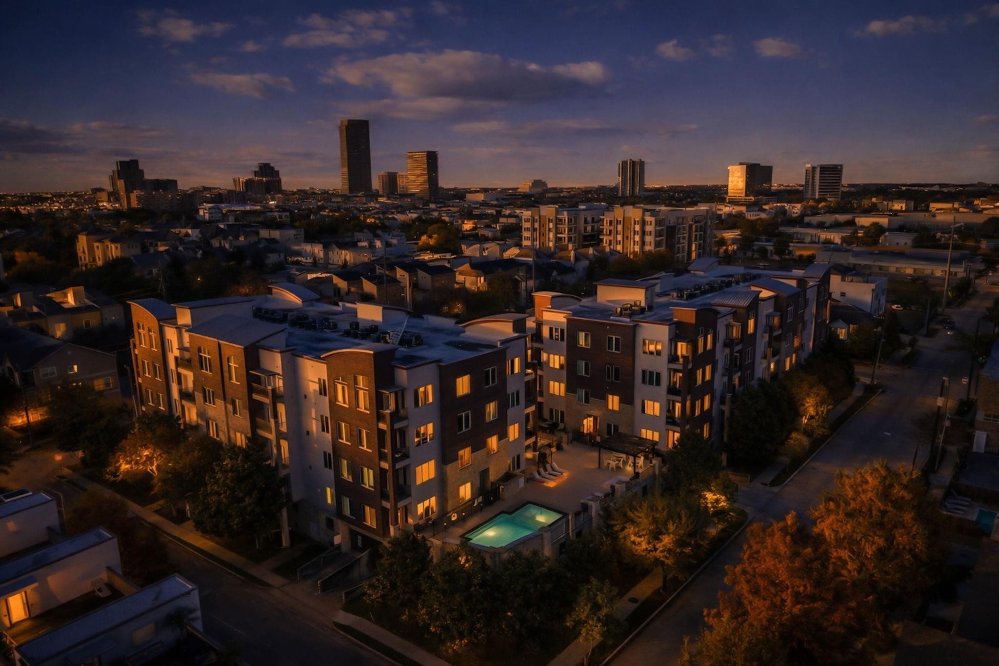1900 Genesee Street, Unit 203 Houston, TX 77006 - Photo 29 of 30 This photo shows a modern apartment complex at dusk, featuring a well-lit exterior and a central swimming pool. It's set in an urban area with a city skyline in the background, offering a blend of residential comfort and city living.