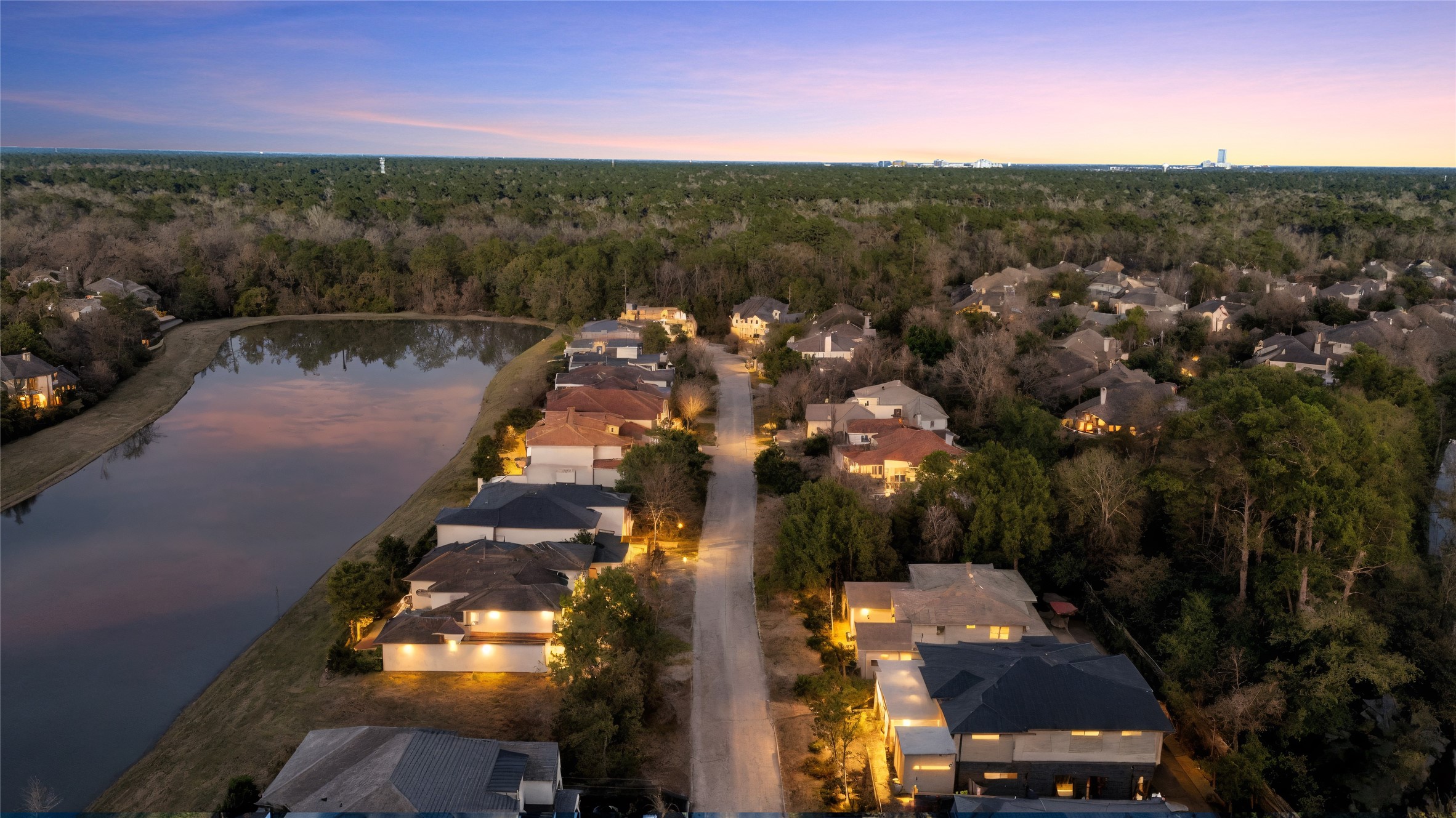 47 Pronghorn Place Spring, TX 77389 - Photo 3 of 48 This aerial photo showcases a serene residential neighborhood at dusk, featuring well-lit homes along a tree-lined street. A peaceful pond reflects the twilight sky, adding to the tranquil suburban setting surrounded by lush greenery.