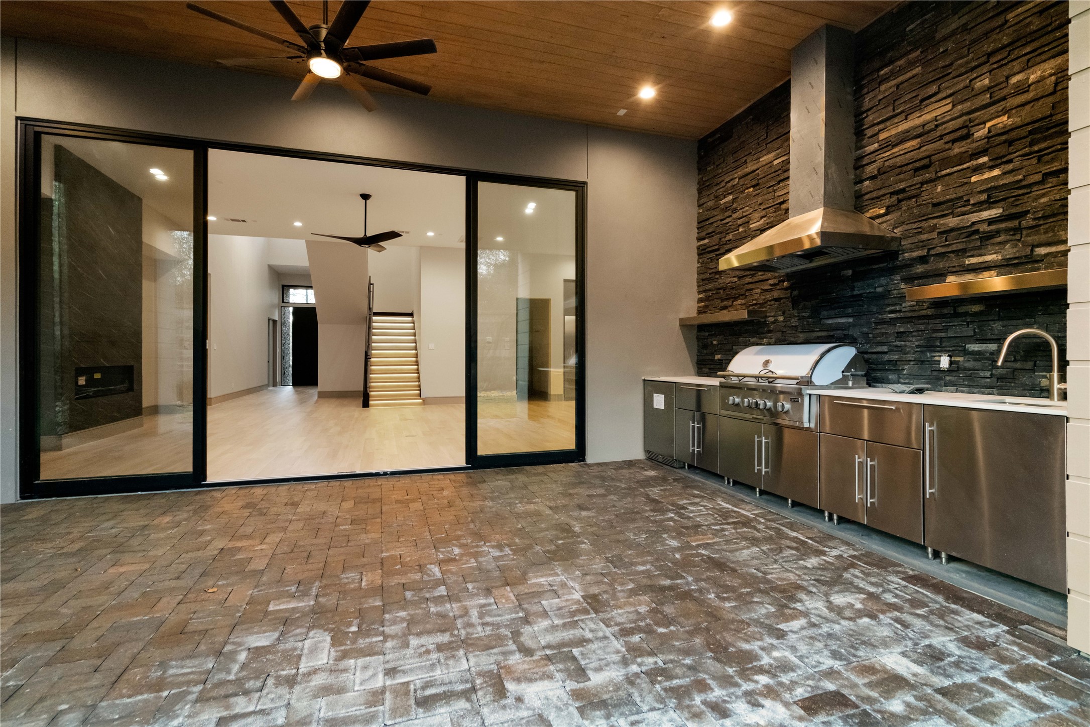 47 Pronghorn Place Spring, TX 77389 - Photo 45 of 48 This photo showcases a modern outdoor kitchen with stainless steel appliances, a built-in grill, and a sink. It features a sleek stone wall and brick flooring, with large sliding glass doors leading to an indoor living space. The area is enhanced by recessed lighting and a ceiling fan, offering a stylish and functional space for entertaining.