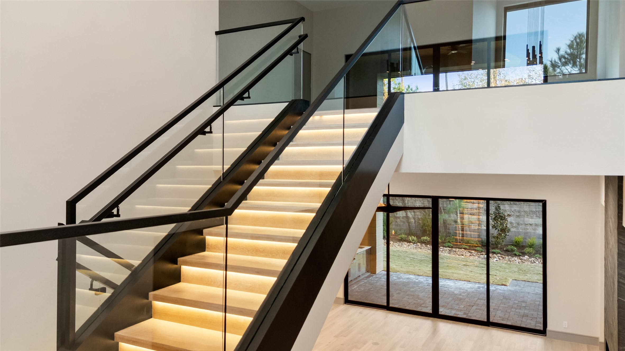 47 Pronghorn Place Spring, TX 77389 - Photo 9 of 48 This modern home features a sleek, illuminated staircase with glass railings and a spacious, brightly lit interior. Large sliding glass doors lead to a landscaped garden, enhancing indoor-outdoor living.