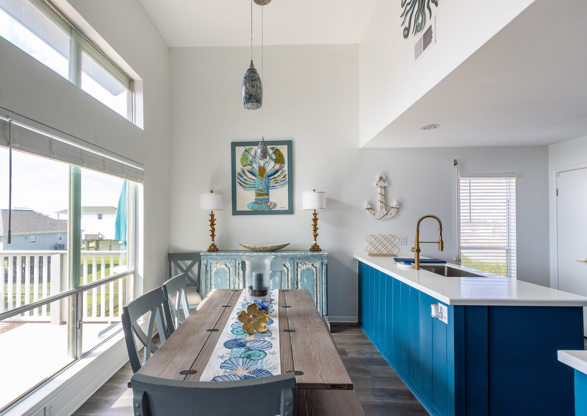 17609 Termini-San Luis Pass Road Galveston, TX 77554 - Photo 27 of 37 This photo showcases a bright, airy dining area with large windows and modern decor. It features a wooden table with stylish chairs, a blue accent cabinet, and nautical-themed artwork. The space seamlessly connects to a sleek kitchen with a white countertop and blue cabinetry, offering a fresh, coastal vibe.