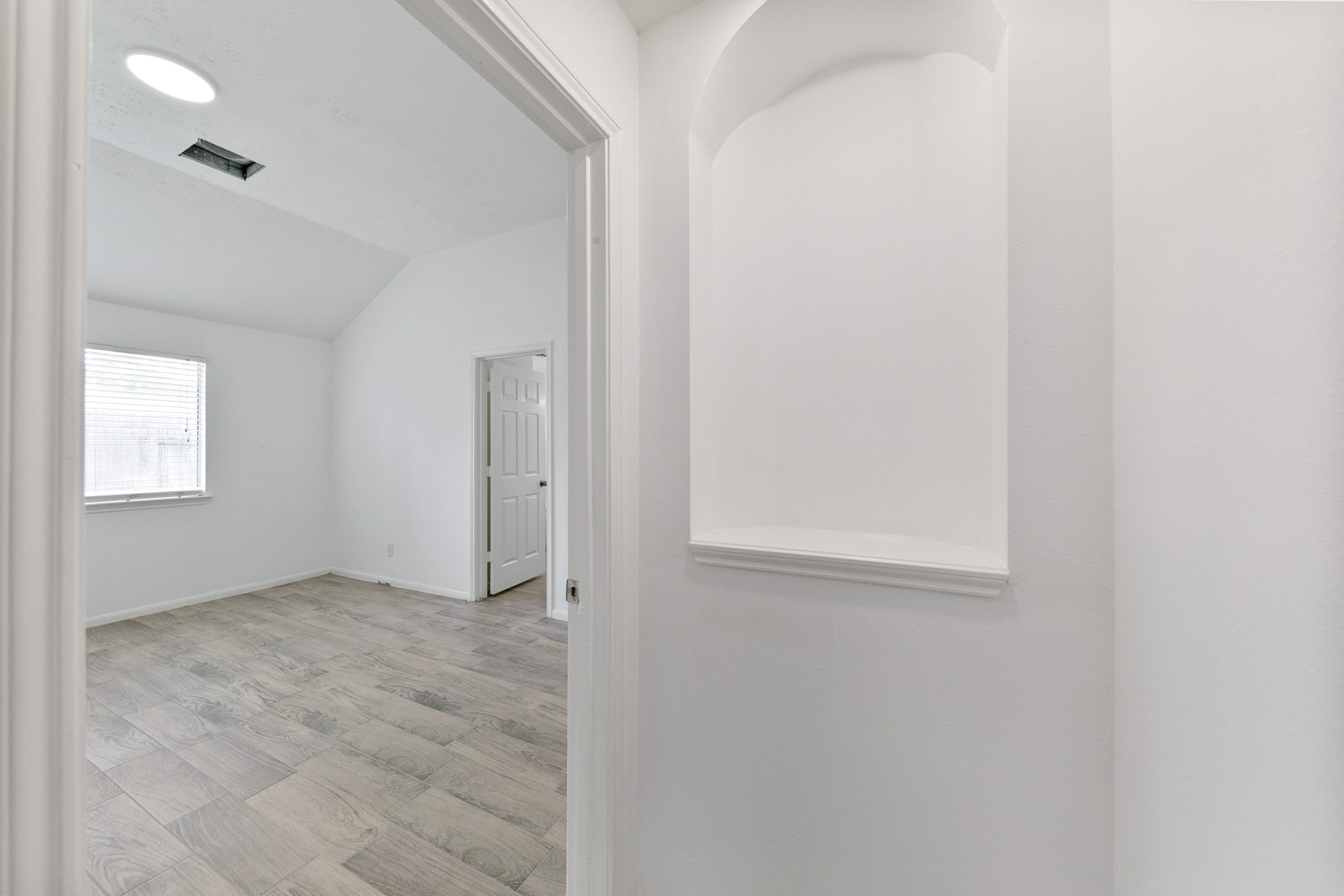 12014 Raven View Drive Houston, TX 77067 - Photo 19 of 29 This photo shows a bright, unfurnished room with light wood flooring, white walls, and a window allowing natural light. There's an open doorway leading to another room and a built-in wall niche for decorative use.