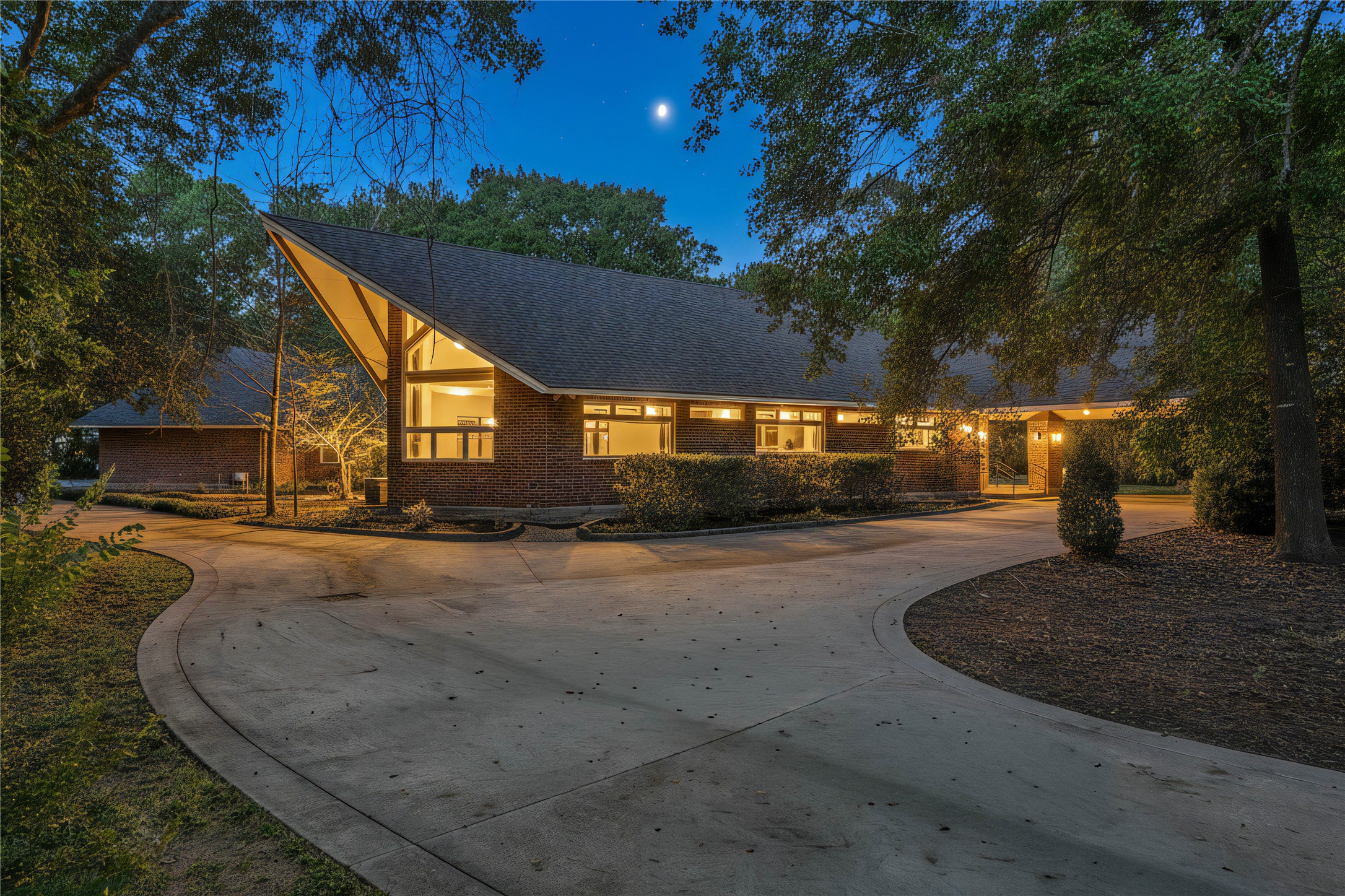16621 Hereford Road Tomball, TX 77377 - Photo 2 of 46 This contemporary gem, with striking architecture, featuring a steep, angular roof and large windows. It's set amidst mature trees, illuminated warmly against a twilight sky, with a spacious curved driveway enhancing its appeal.