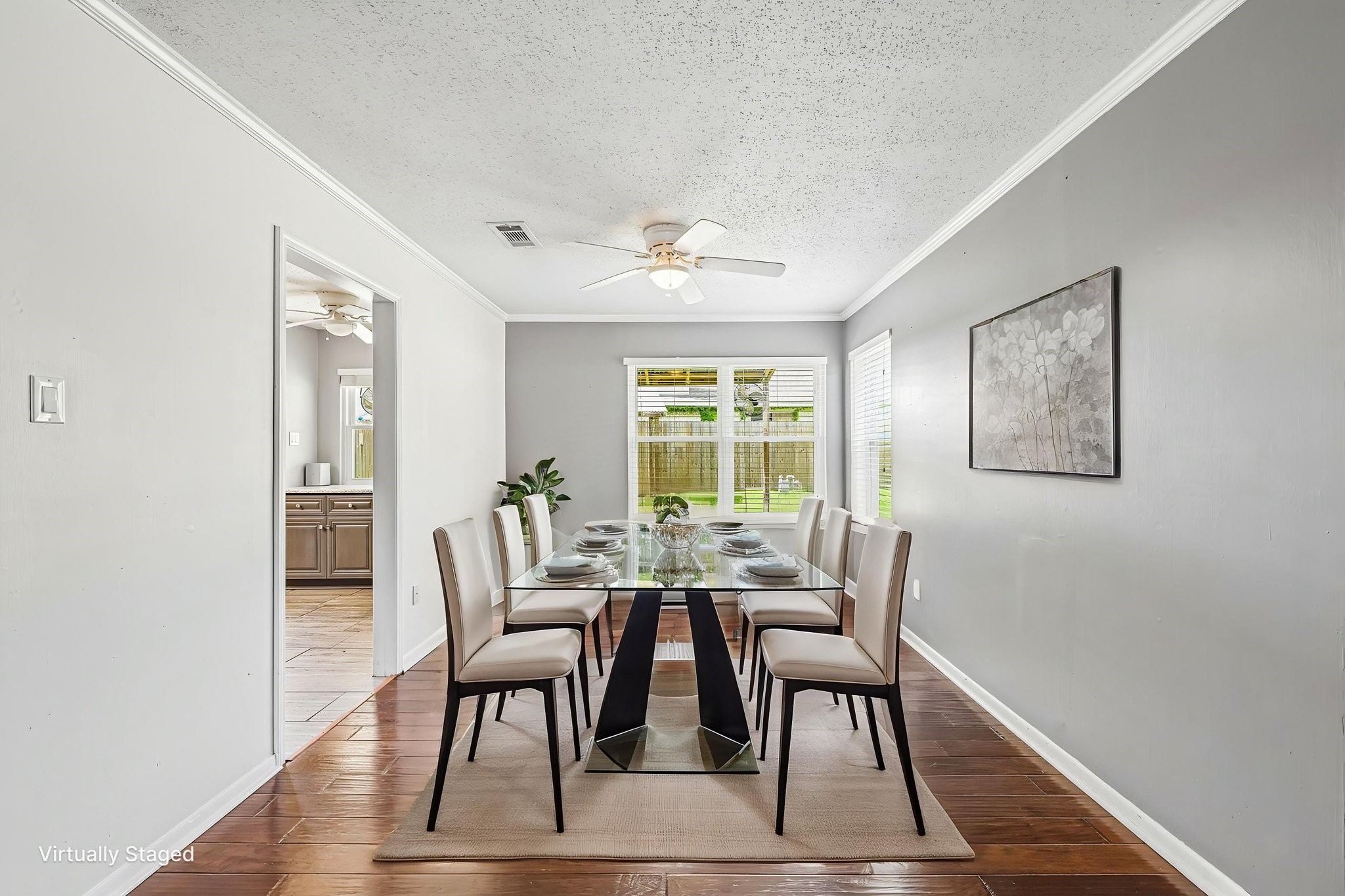 8 Arlington Street Conroe, TX 77301 - Photo 3 of 37 This photo shows a modern dining room with a glass table set for six. It features hardwood floors, gray walls, white trim, and a ceiling fan. Large windows provide ample natural light, with a view of the backyard. The space connects to the kitchen through an open doorway.