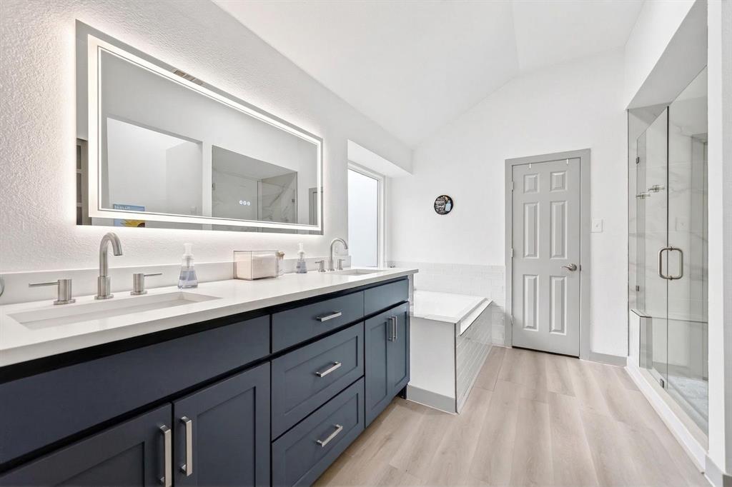 7503 Vista Ridge Lane Sachse, TX 75048 - Photo 20 of 33 Modern bathroom featuring a dual vanity with dark cabinetry and light countertops, a rectangular mirror with backlighting, a built-in bathtub, and a glass-enclosed shower