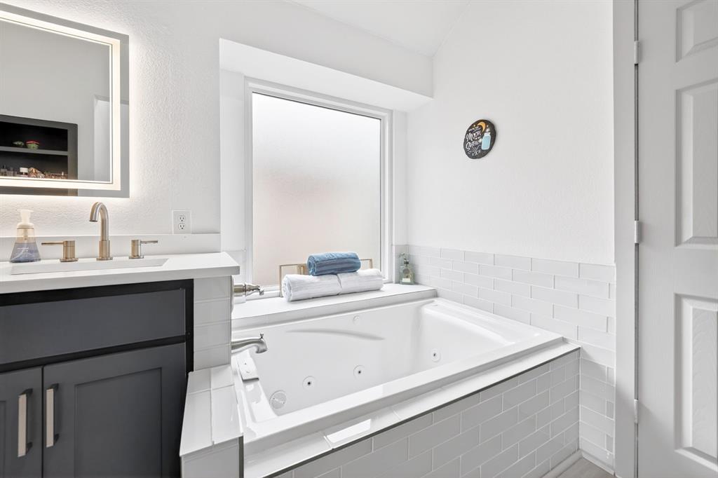 7503 Vista Ridge Lane Sachse, TX 75048 - Photo 22 of 33 The bathroom features a built-in bathtub with white tile surround, a window, and a vanity with a rectangular sink, brushed nickel faucet, and an illuminated mirror