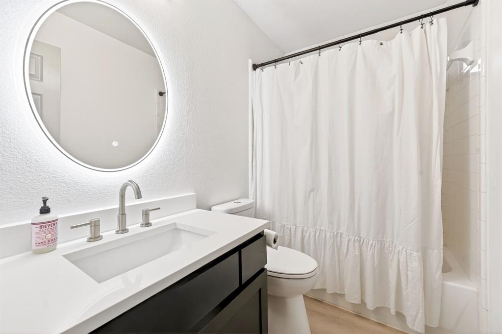 7503 Vista Ridge Lane Sachse, TX 75048 - Photo 26 of 33 The bathroom features a modern vanity with a white countertop and dark cabinetry, a back-lit round mirror, and a shower with a white curtain